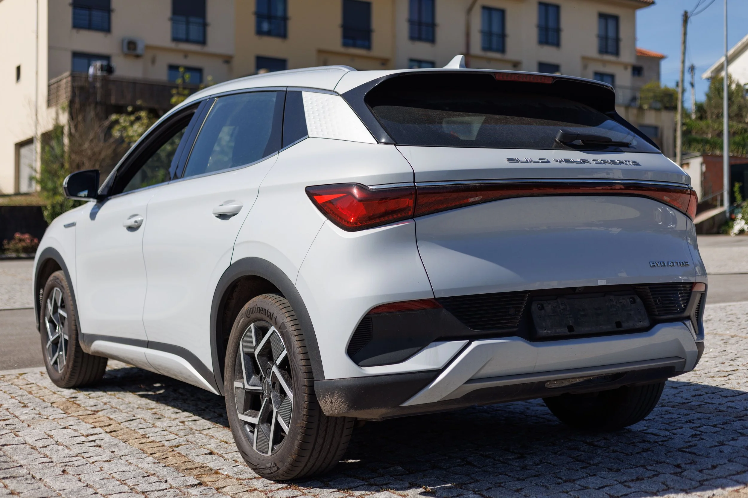 Carro branco estacionado em calçada de pedras, com edifícios residenciais ao fundo.