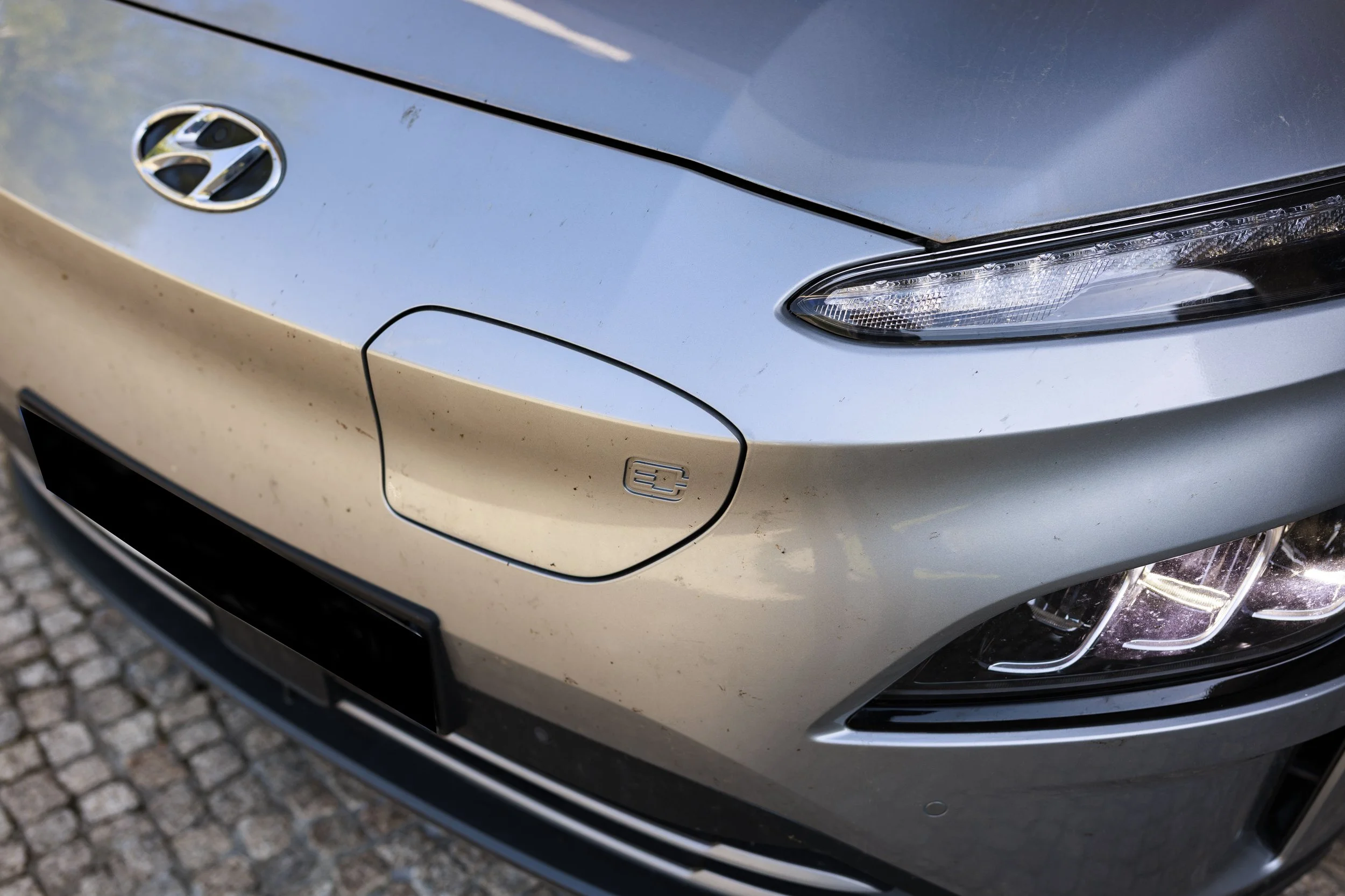 Parte frontal de um carro prata com detalhes em preto, incluindo faróis, grade frontal, logo da Hyundai e símbolo de combustível, estacionado em rua de calçamento de pedras.