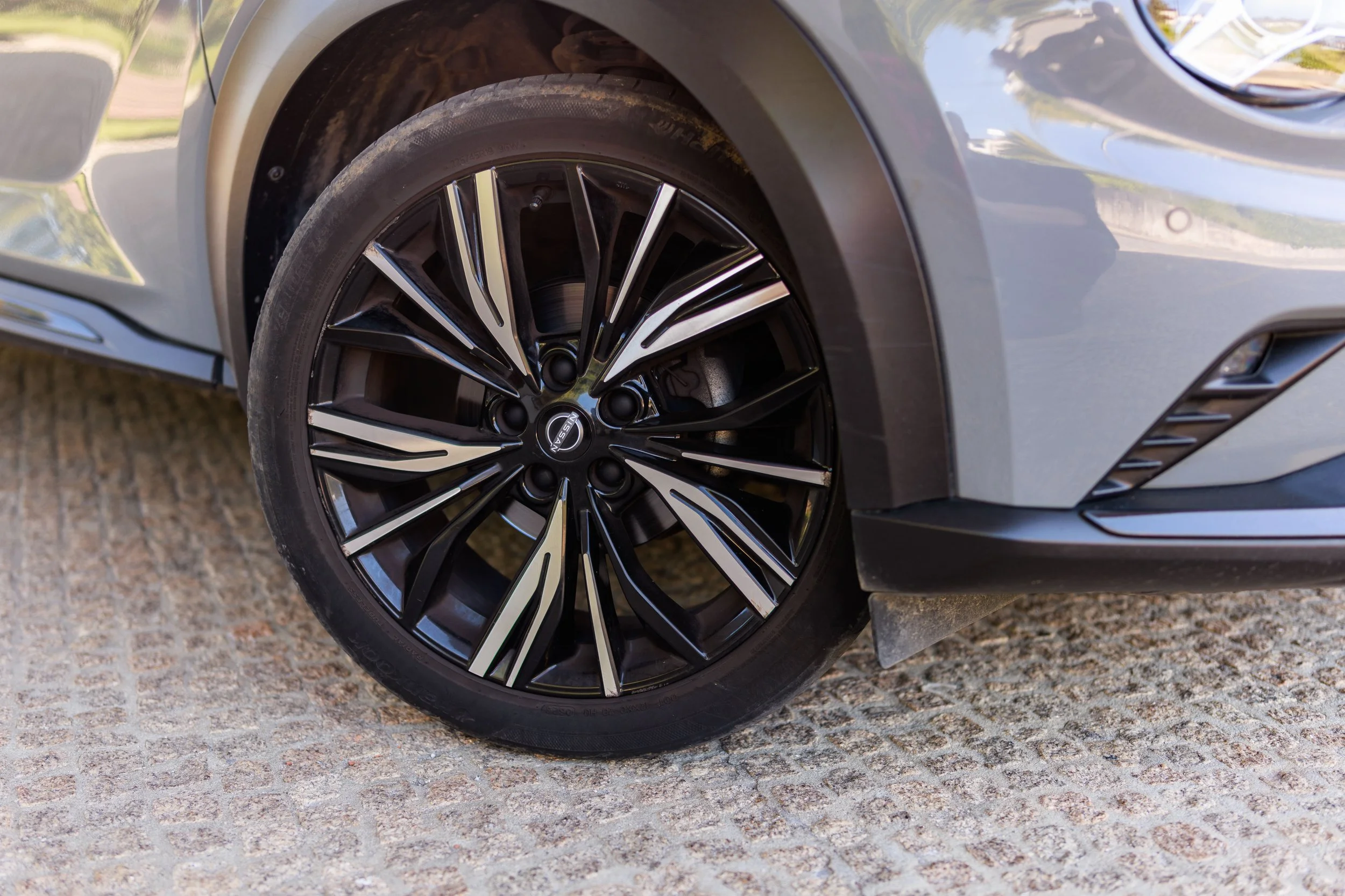 Pneu de carro preto com detalhes prateados e roda de liga leve, parte do parafuso e da roda visíveis, o carro está estacionado sobre uma superfície de pedras.