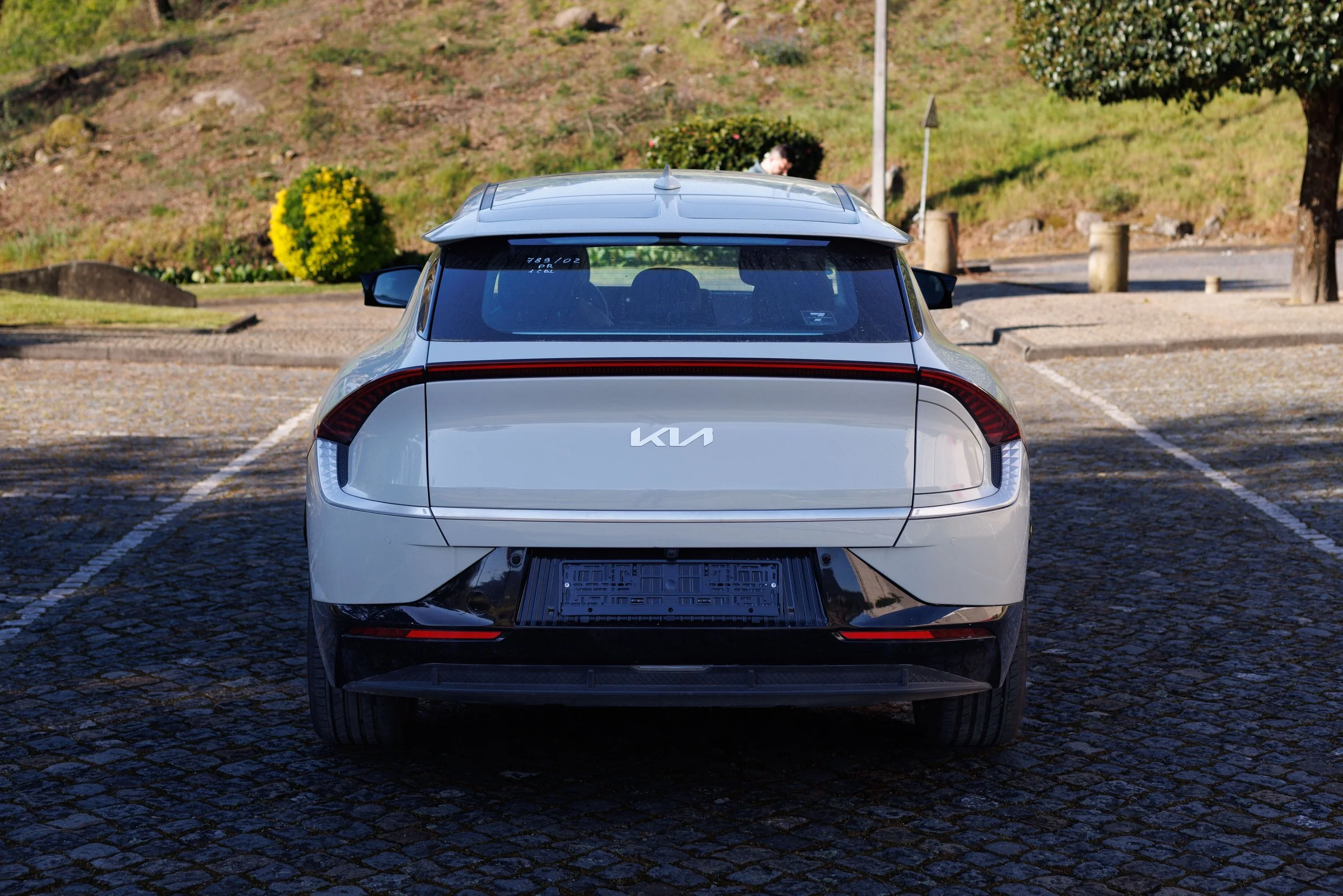 Carro branco da marca Kia estacionado em uma vaga de estacionamento de calçamento de pedras, ao fundo há gramado, árvores, arbustos floridos e uma lixeira na calçada.