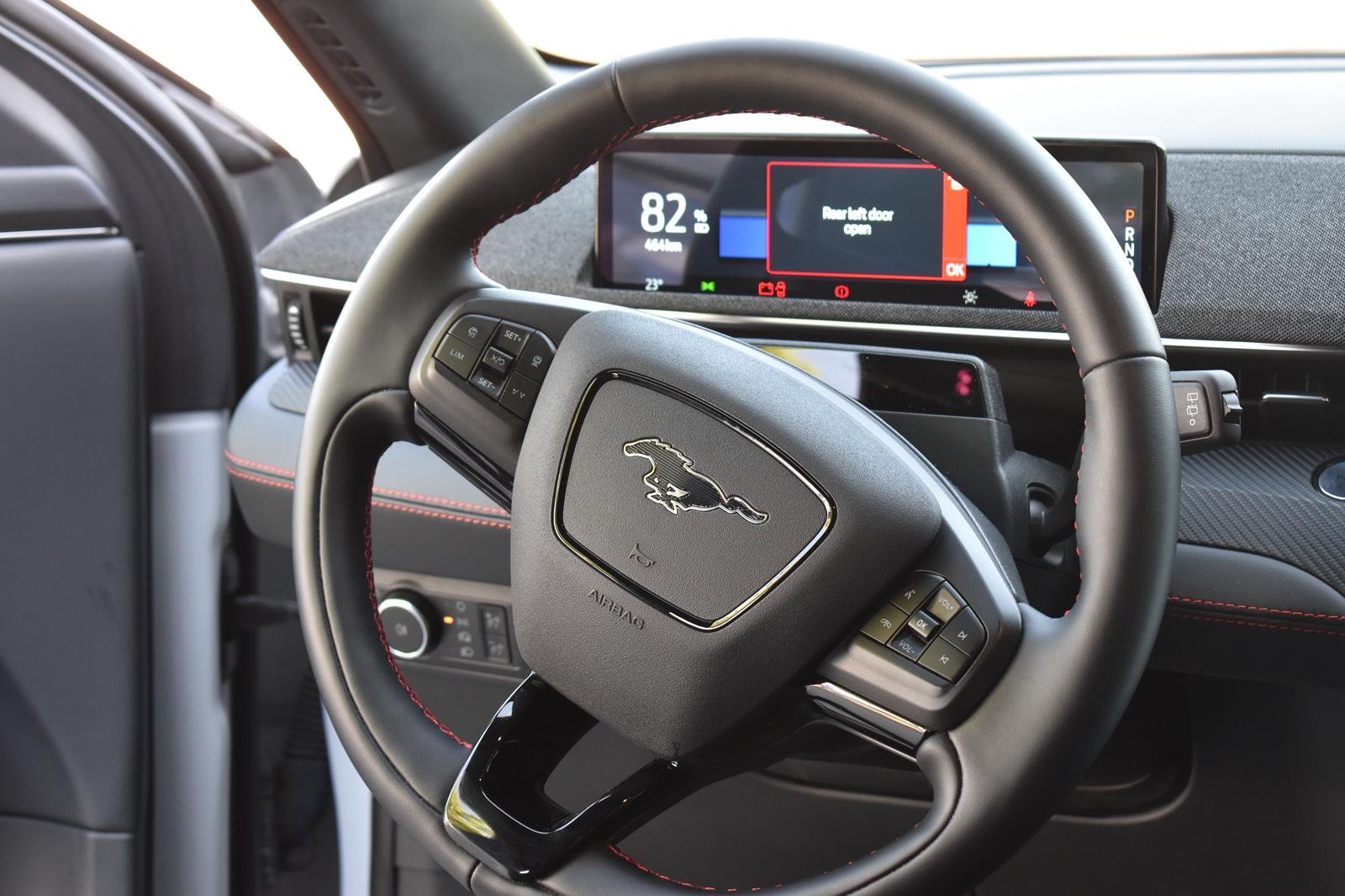 Painel digital do carro exibindo aviso de porta traseira aberta, ao fundo o volante com logo do Ford Mustang adaptado ao estilo do carro Ford, com controles do lado esquerdo e direito.