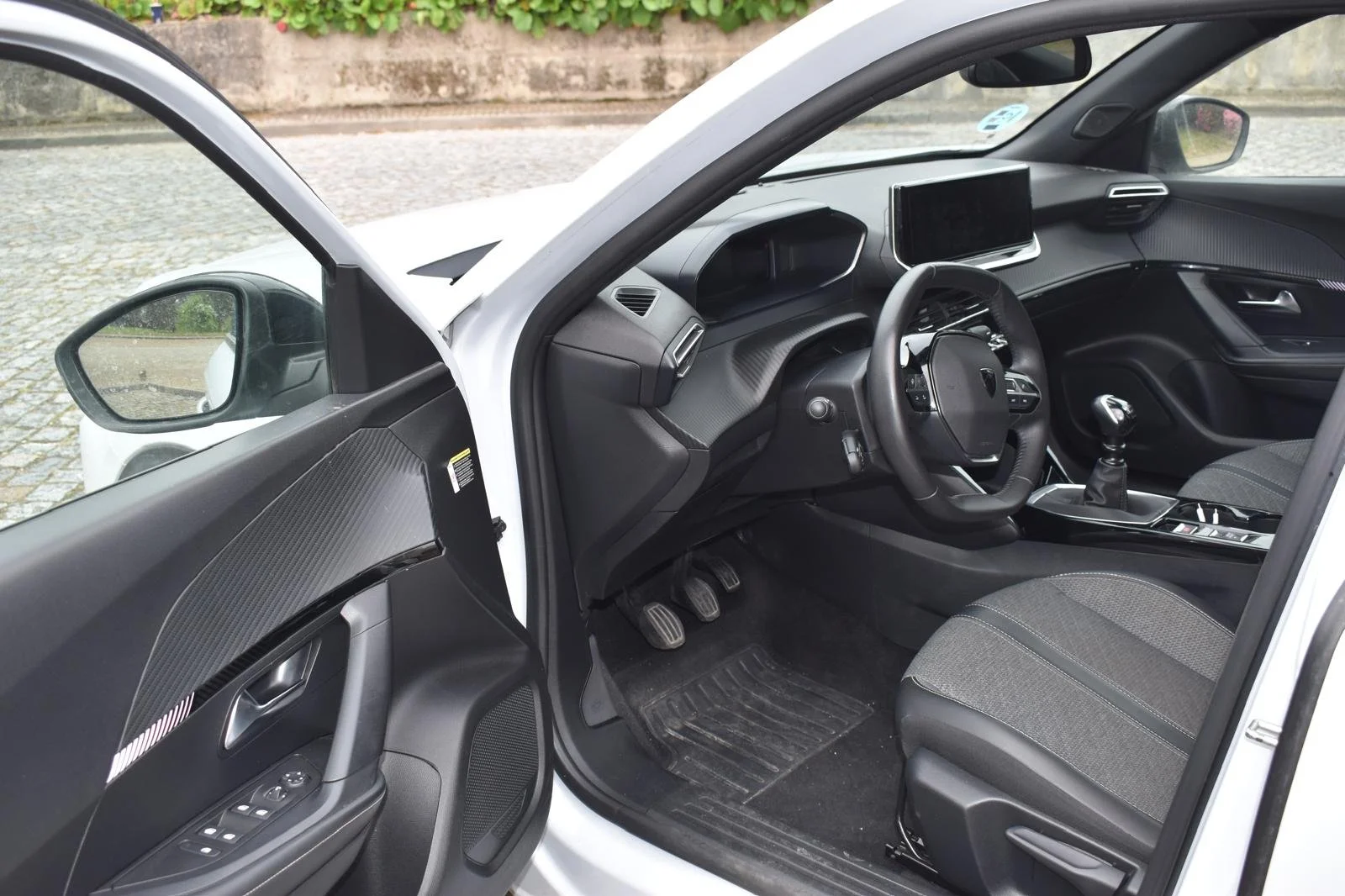Interior de um carro branco, mostrando o painel, volante, banco e pedais.