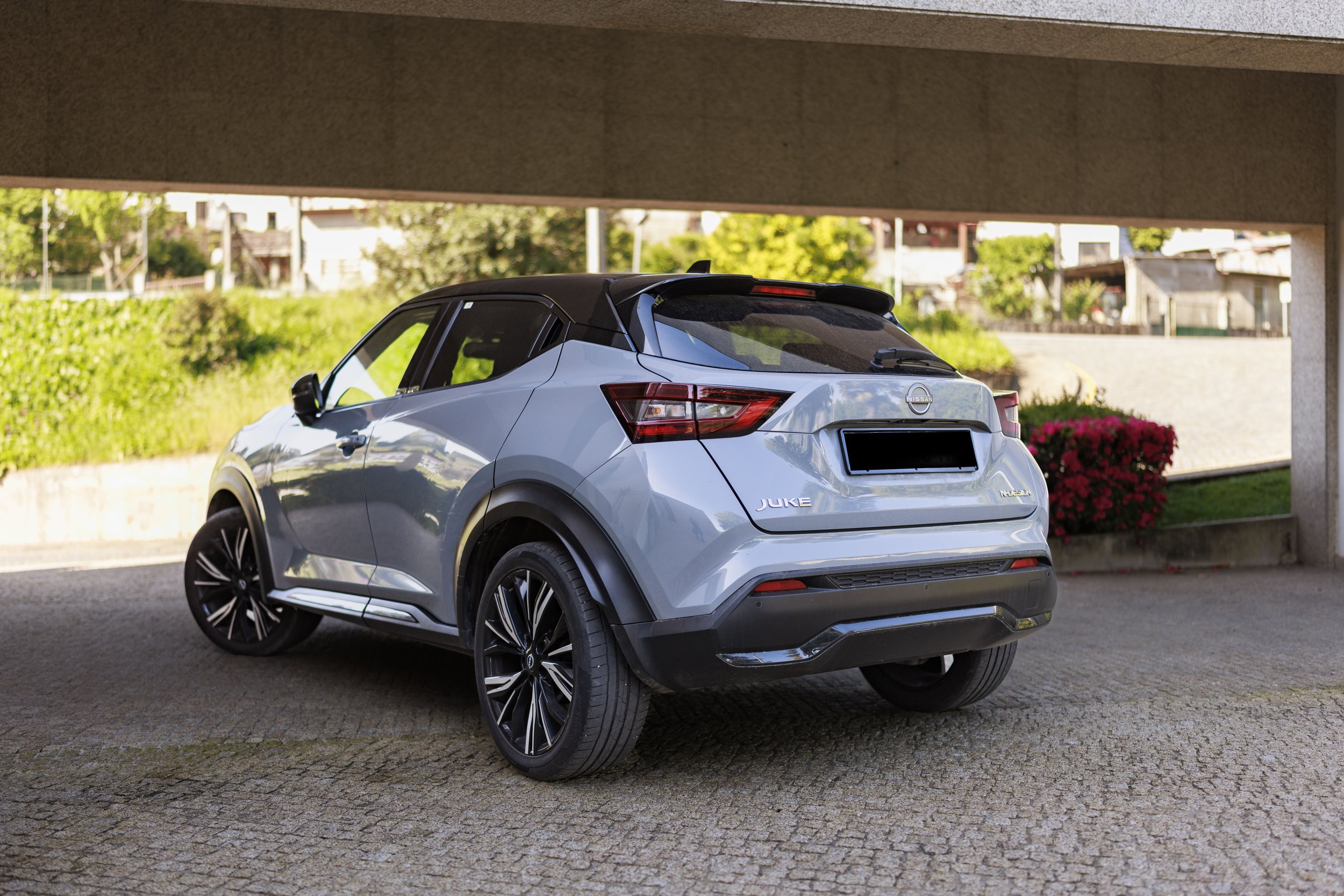 Carro Nissan Juke cinza estacionado sob uma cobertura em uma área urbana com árvores, casas e plantas ao fundo.