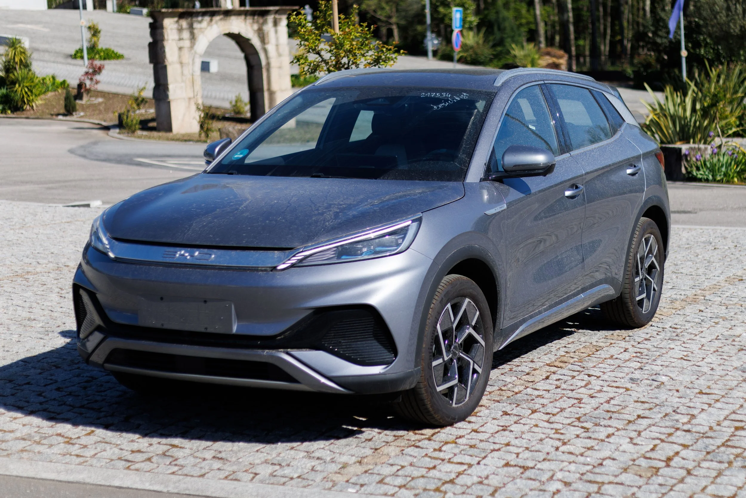 Carro prateado estacionado numa praça com calçada de pedra, árvores e vegetação ao fundo.