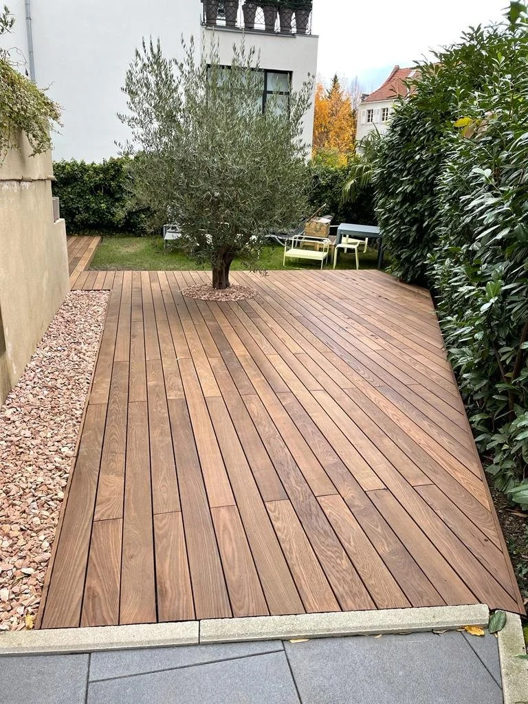 Une terrasse en bois avec un arbre au centre, entourée de haies greenery et d'une façade d'immeuble en arrière-plan.