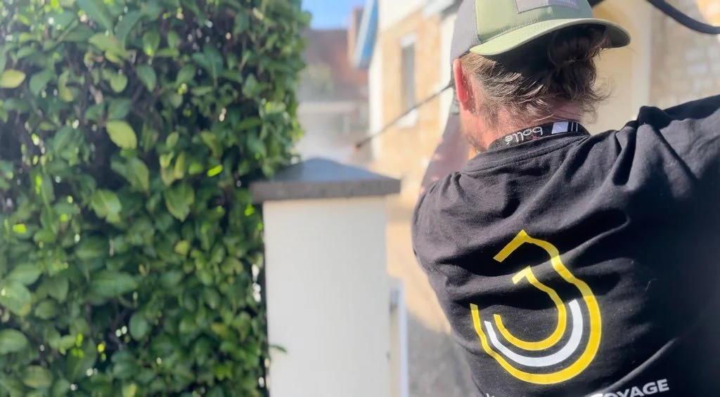 Une personne porte un t-shirt noir avec un logo jaune et gris, et un chapeau gris, visibles de dos, qui travaille à l'extérieur près d'une haie verte.