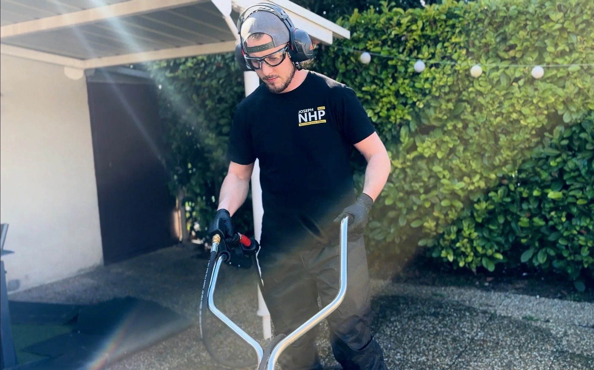 Homme portant un casque antibruit, lunettes de protection, gants et t-shirt noir avec le logo 'JOSEPH NHP', utilisant une pioche électrique dans un espace extérieur avec des buissons verts en arrière-plan.