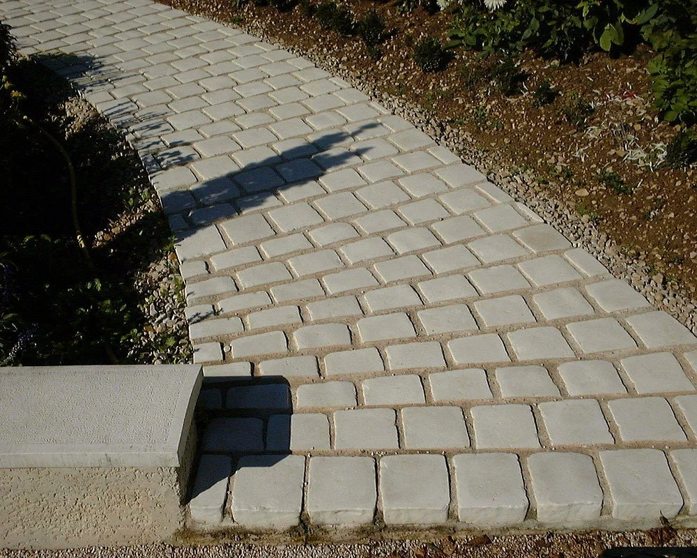 Chemin pavé en pierre avec des buissons à droite et une clôture ou un rebord en béton en bas à gauche.