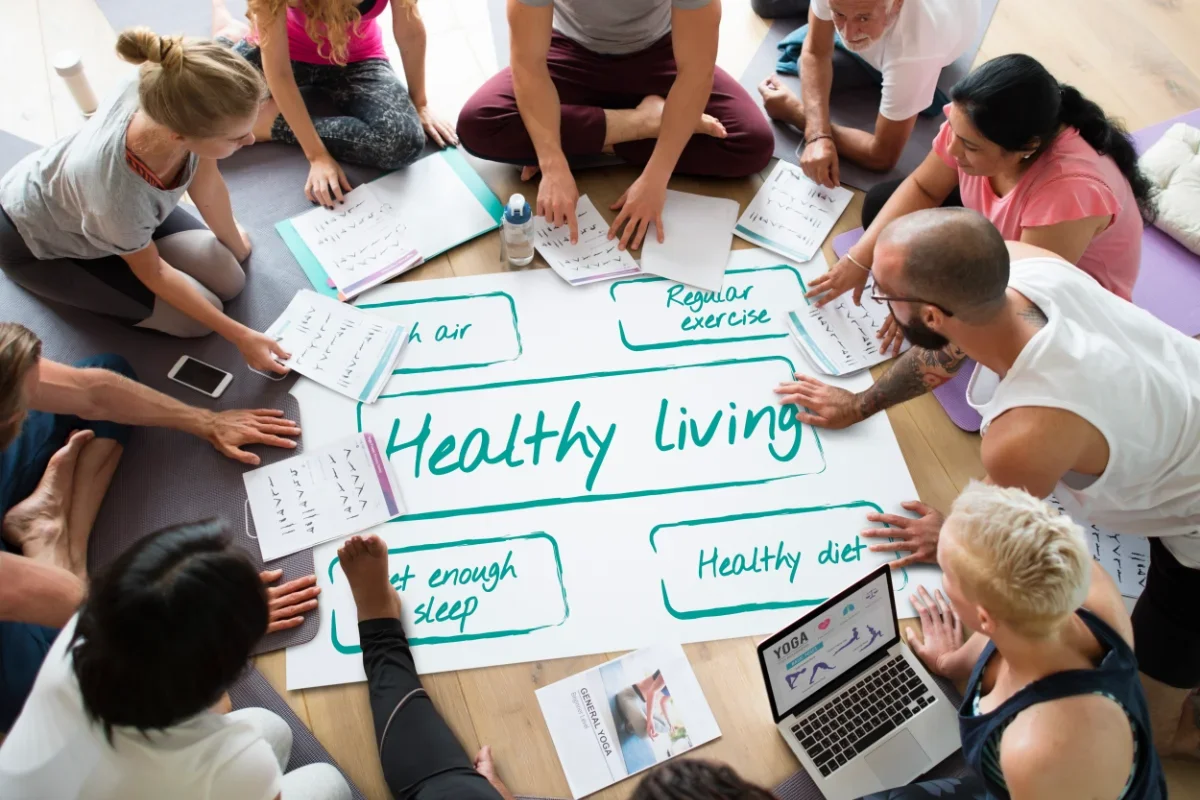 Group of people sitting around a table discussing ideas for healthy living, with notes on topics such as sleep, exercise, and diet written on a large sheet of paper.