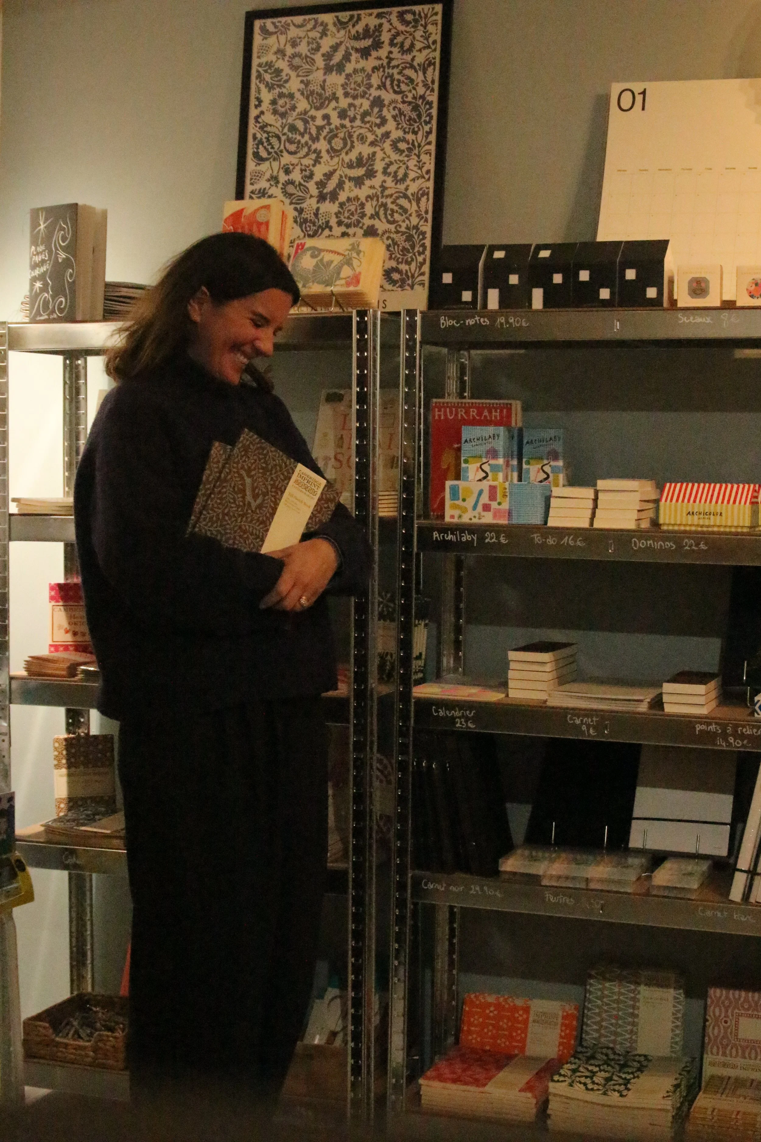 A woman with dark hair smiling and holding several books, standing in front of metal shelves filled with books, notebooks, and decorative items in a bookstore or library.