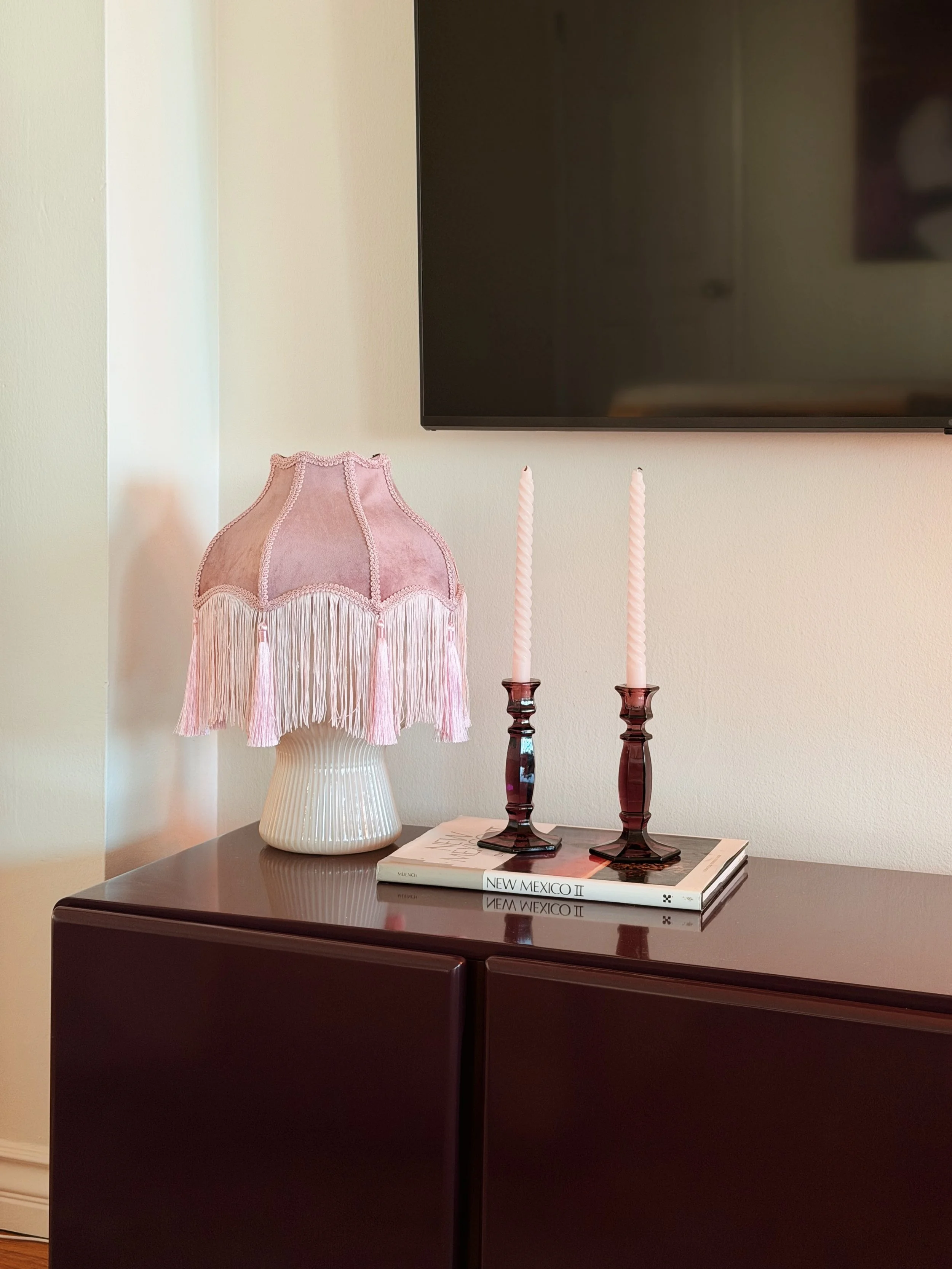 A dark wooden sideboard with a pink fringed table lamp, two pink and purple candlesticks, and a stack of magazines, with a mounted TV and a wall in the background.