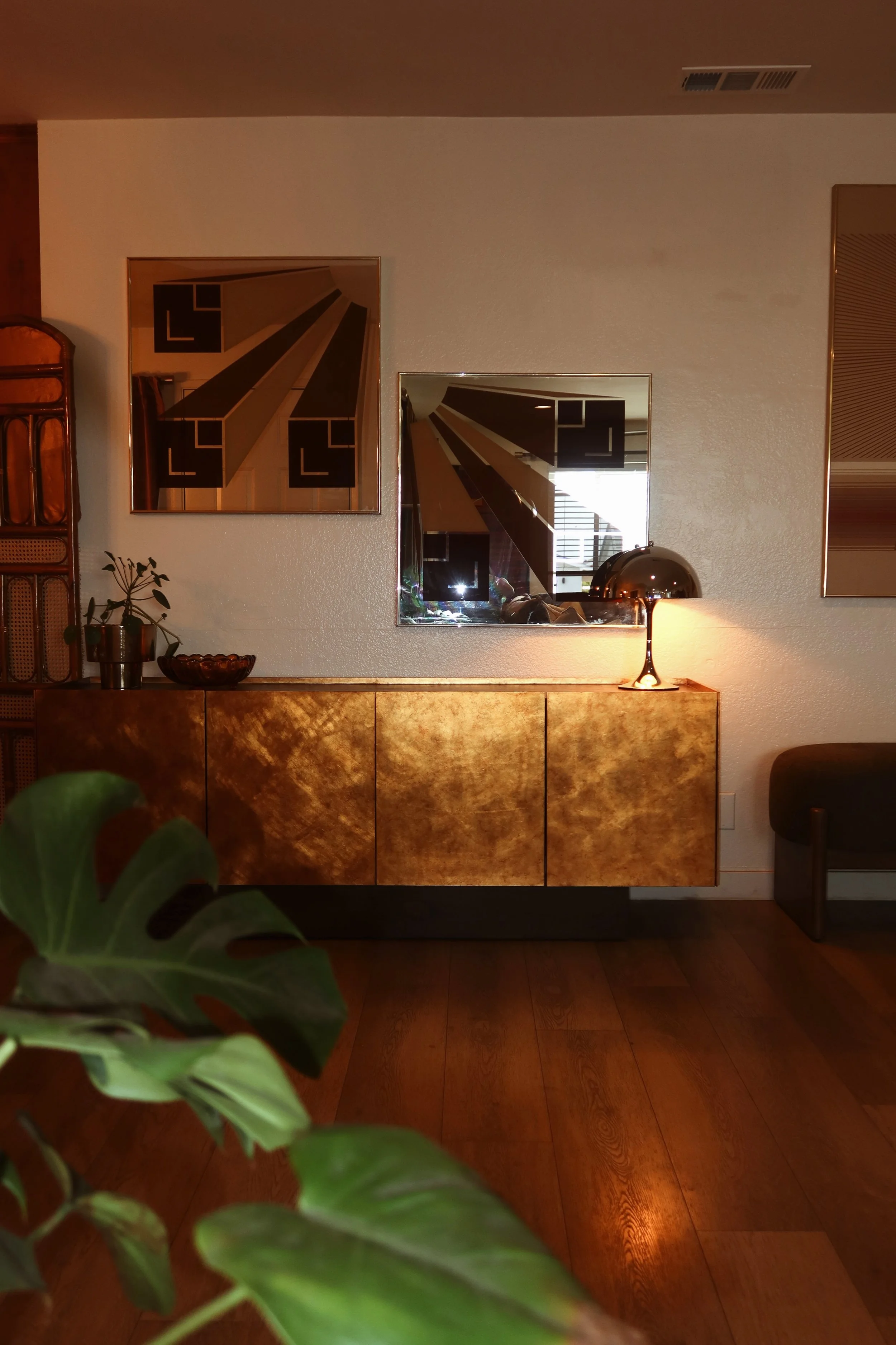 A vintage wooden sideboard with a gold finish, a potted plant, and a decorative bowl on top sits against a textured white wall. Two geometric paintings and a modern table lamp are also visible.