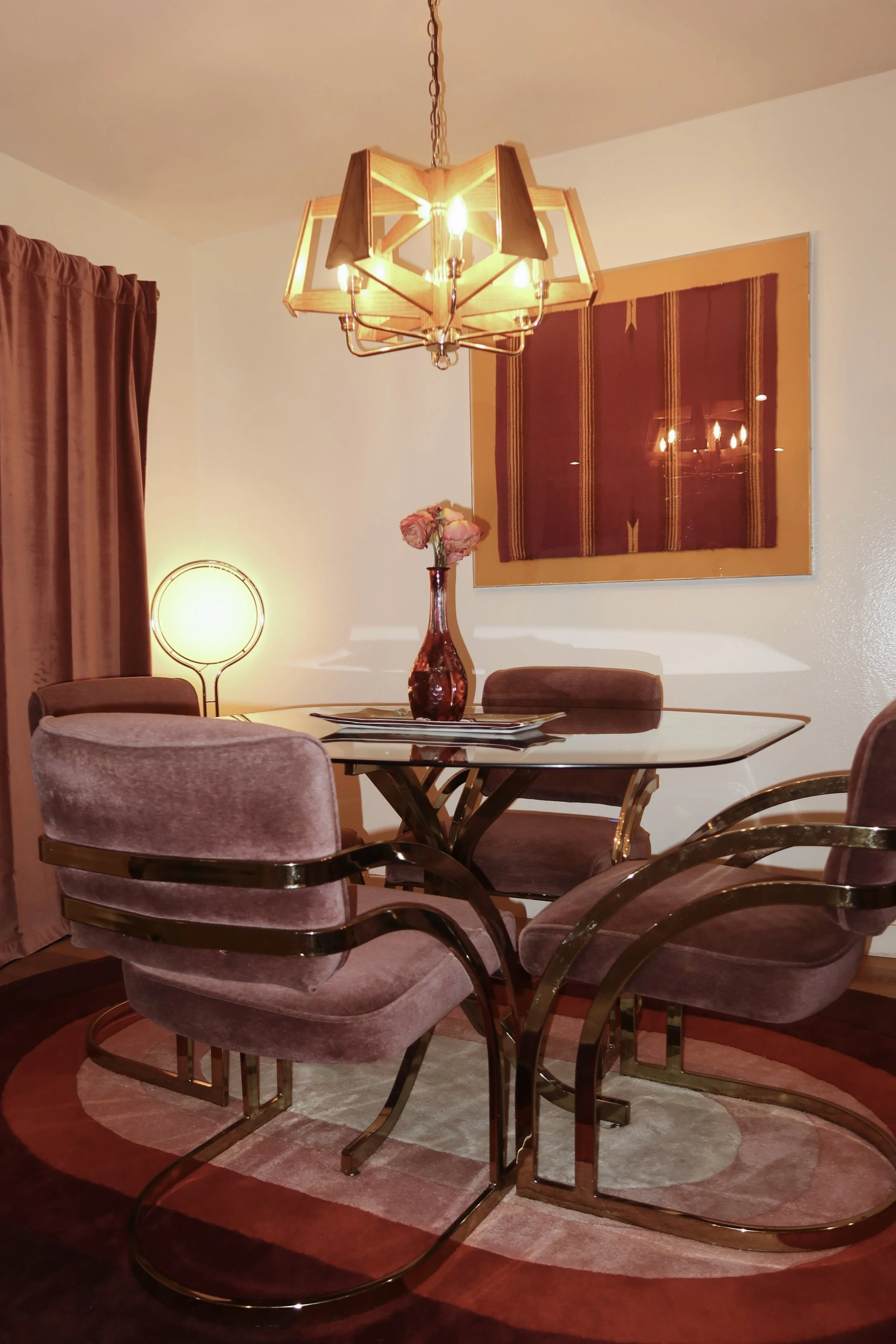 A dining room with a glass table surrounded by velvet pink chairs with brass armrests. A decorative rug under the table, a urn with flowers, a wall hanging, and two lamps, one on each side of the room. A chandelier hangs above the table.