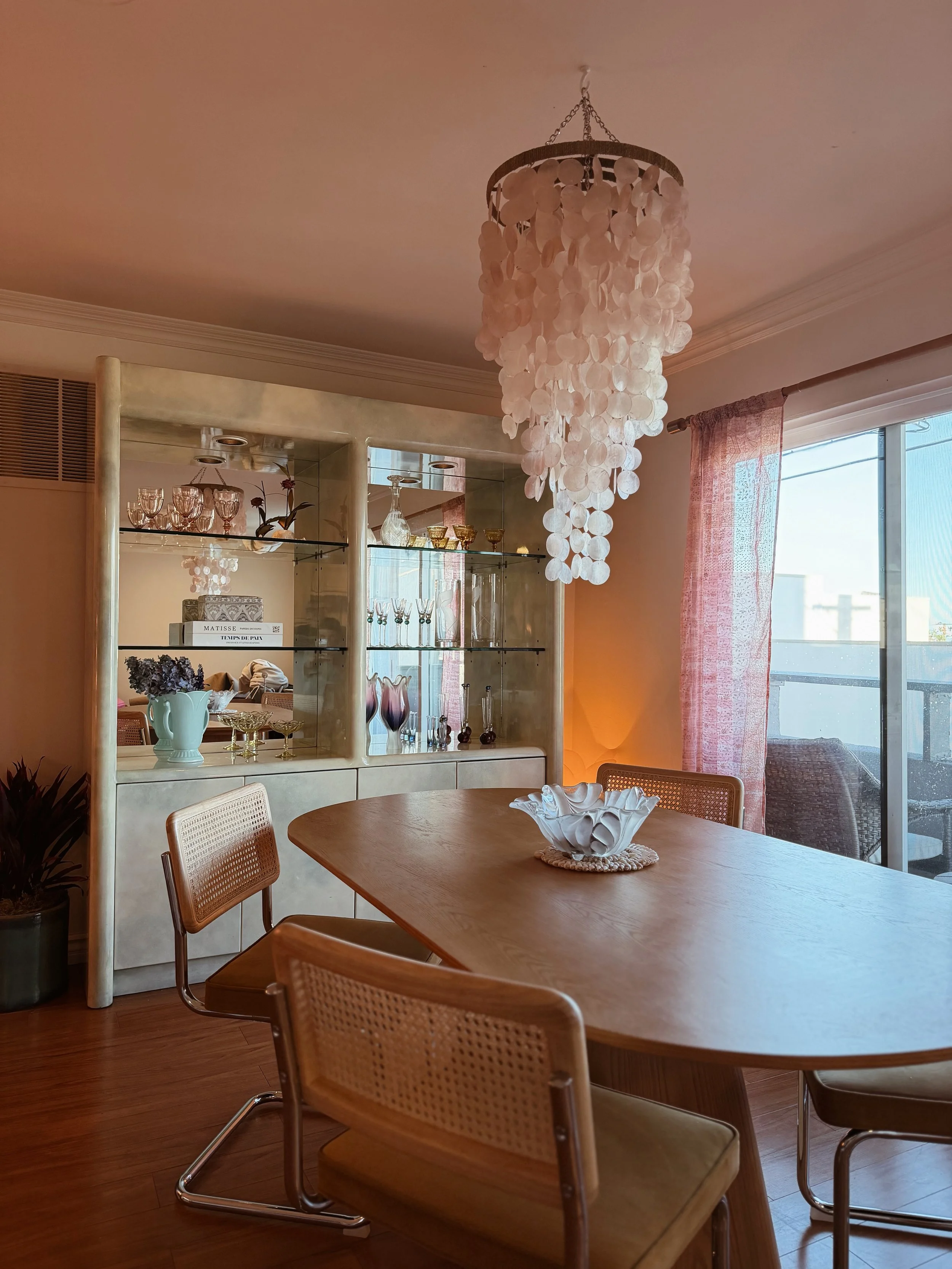 Interior view of a dining room with a wooden table, beige chairs, a chandelier, a glass display cabinet with various glassware and decorative items, a pink curtain, and a large window letting in natural light.