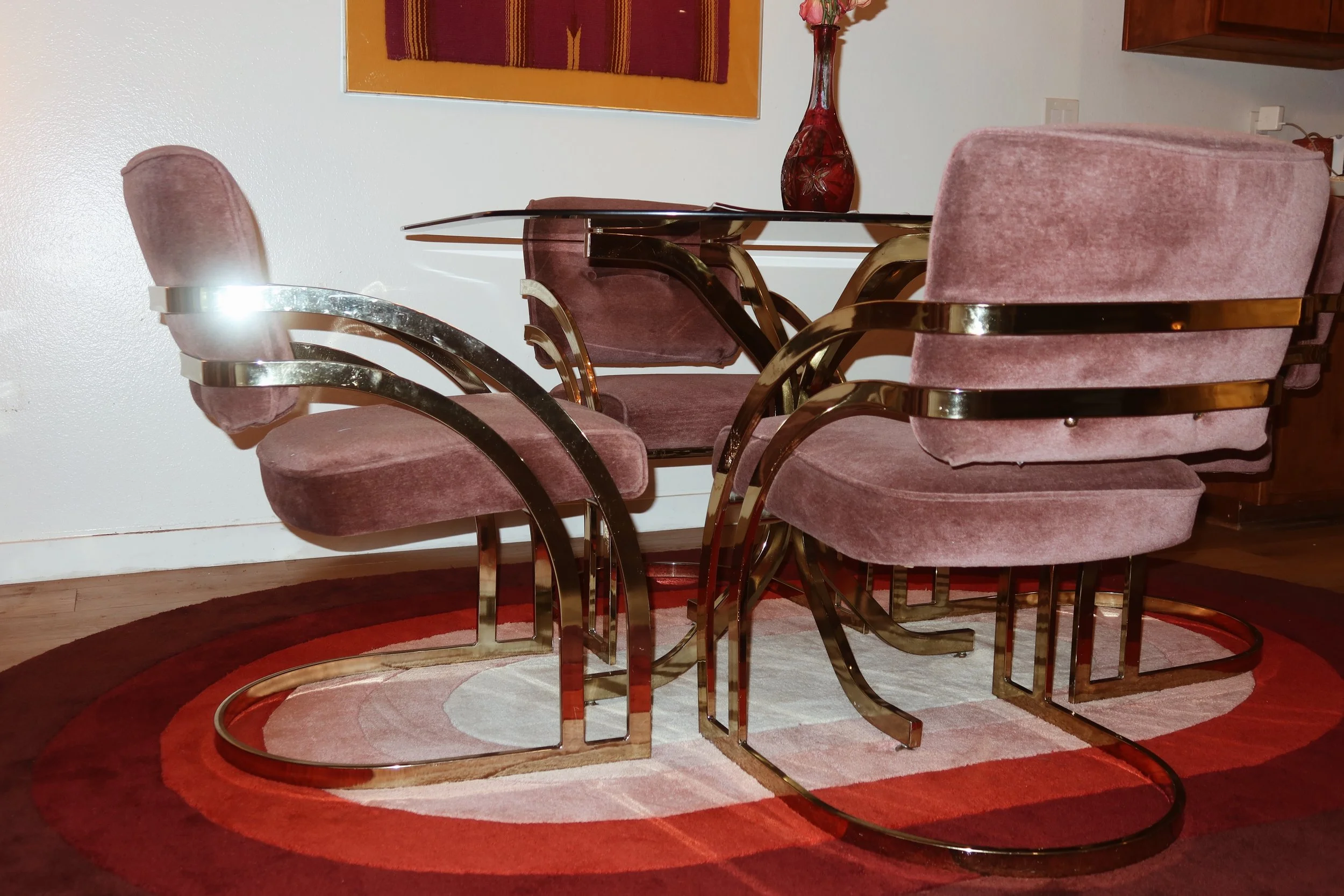 A round dining table with five pink velvet chairs with gold metal arms and legs, placed on a circular red and beige rug in a room with hardwood flooring. A red vase with flowers and a wall hanging are visible on the wall behind the table.