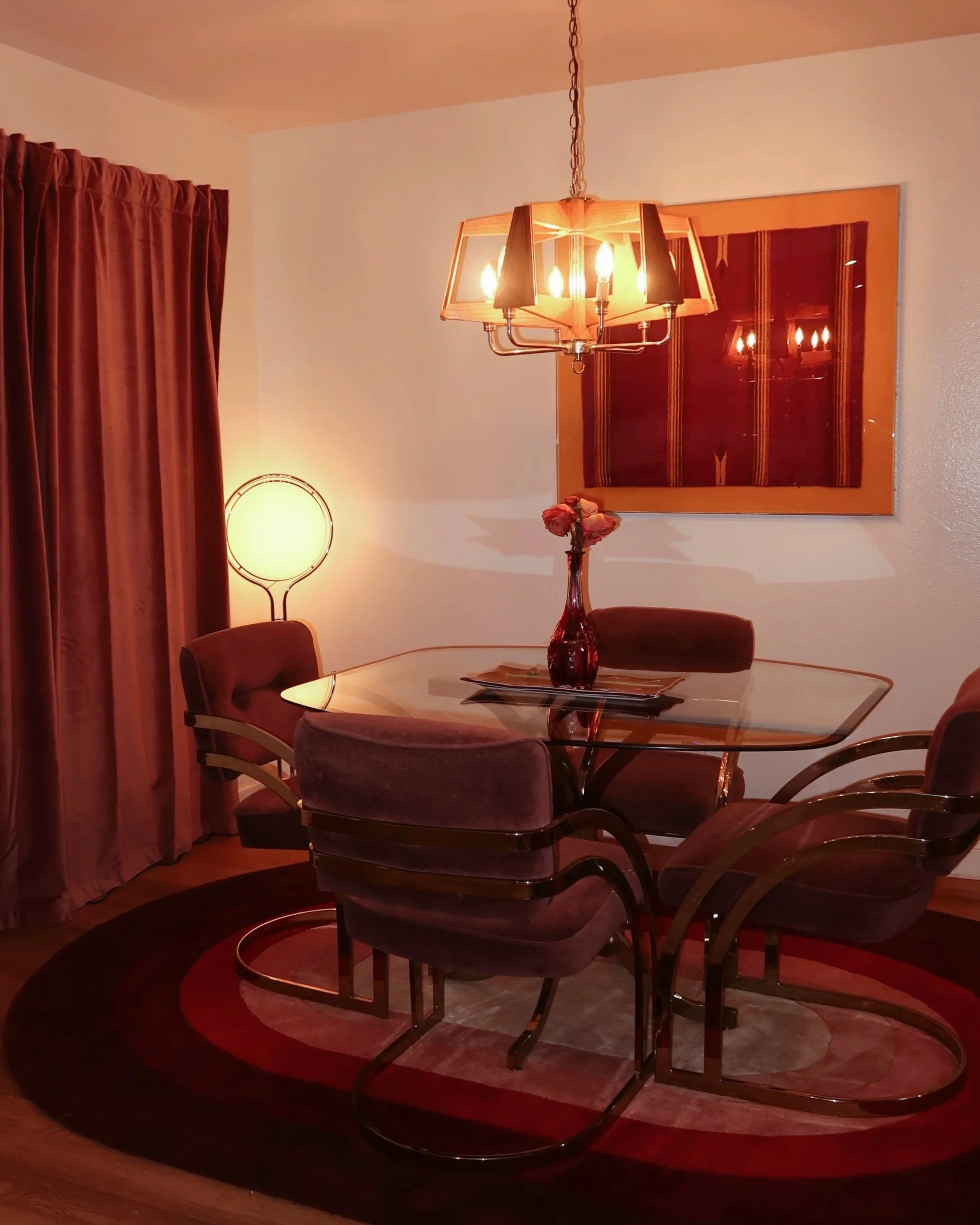 A dining room with a round glass table, surrounded by four chairs with purple velvet seats and armrests. There is a floral centerpiece on the table. A large, wooden framed wall hanging with a dark red and gold pattern, a tall floor lamp with a circul