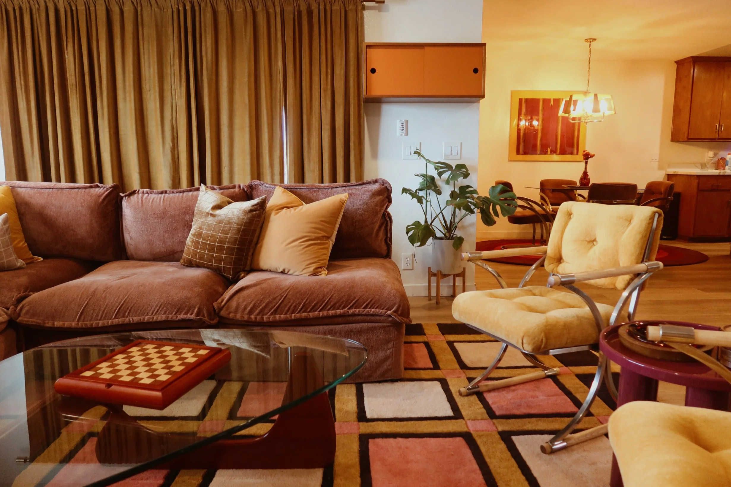 A living room with a large brown sofa with multiple cushions, a glass coffee table with a chessboard, a patterned rug, a yellow armchair, a potted plant, and a dining area with a wooden table, chairs, and a chandelier. Gold curtains cover the window.