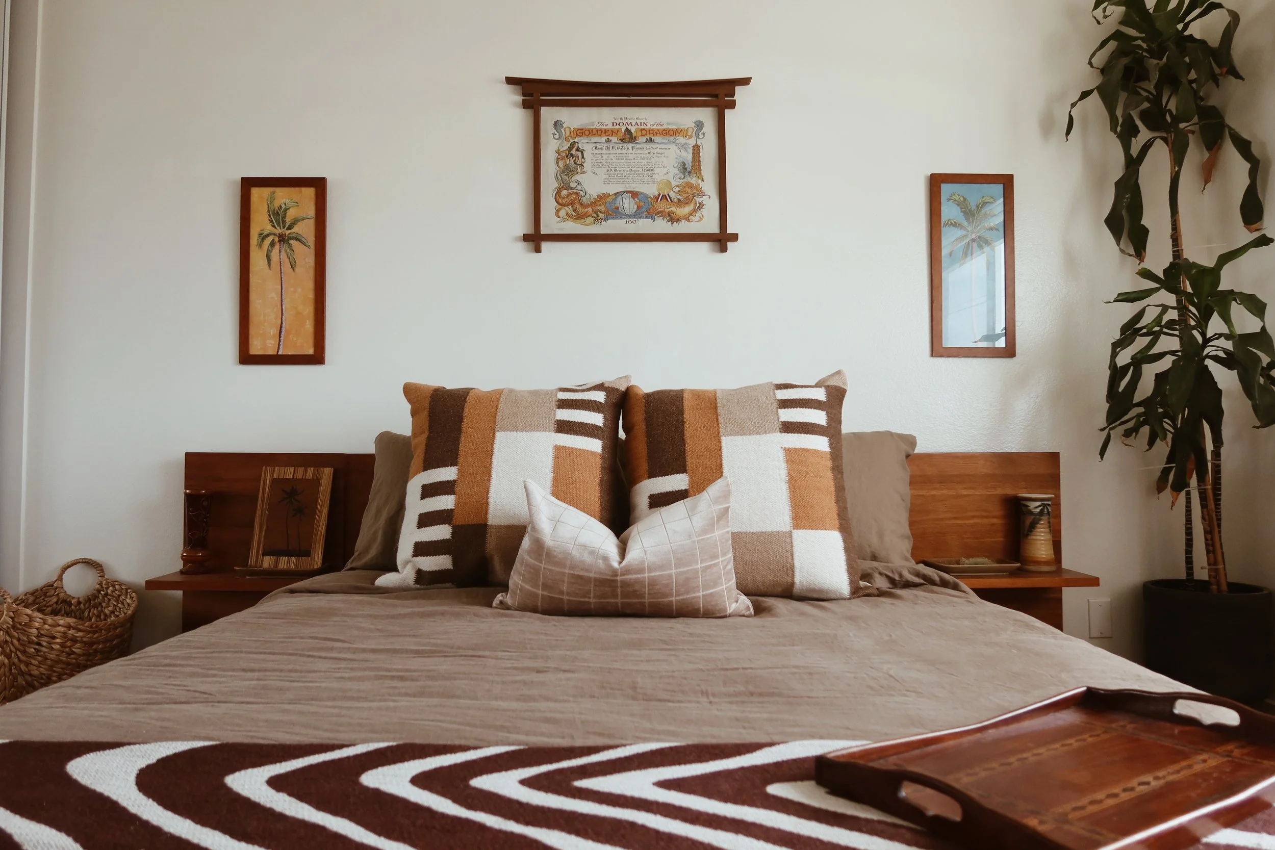 A cozy bedroom features a bed with patterned pillows, a wooden headboard, and framed artwork of palm trees and a dragon map on the wall. A large potted plant is on the right side, and a basket is on the left side of the bed.