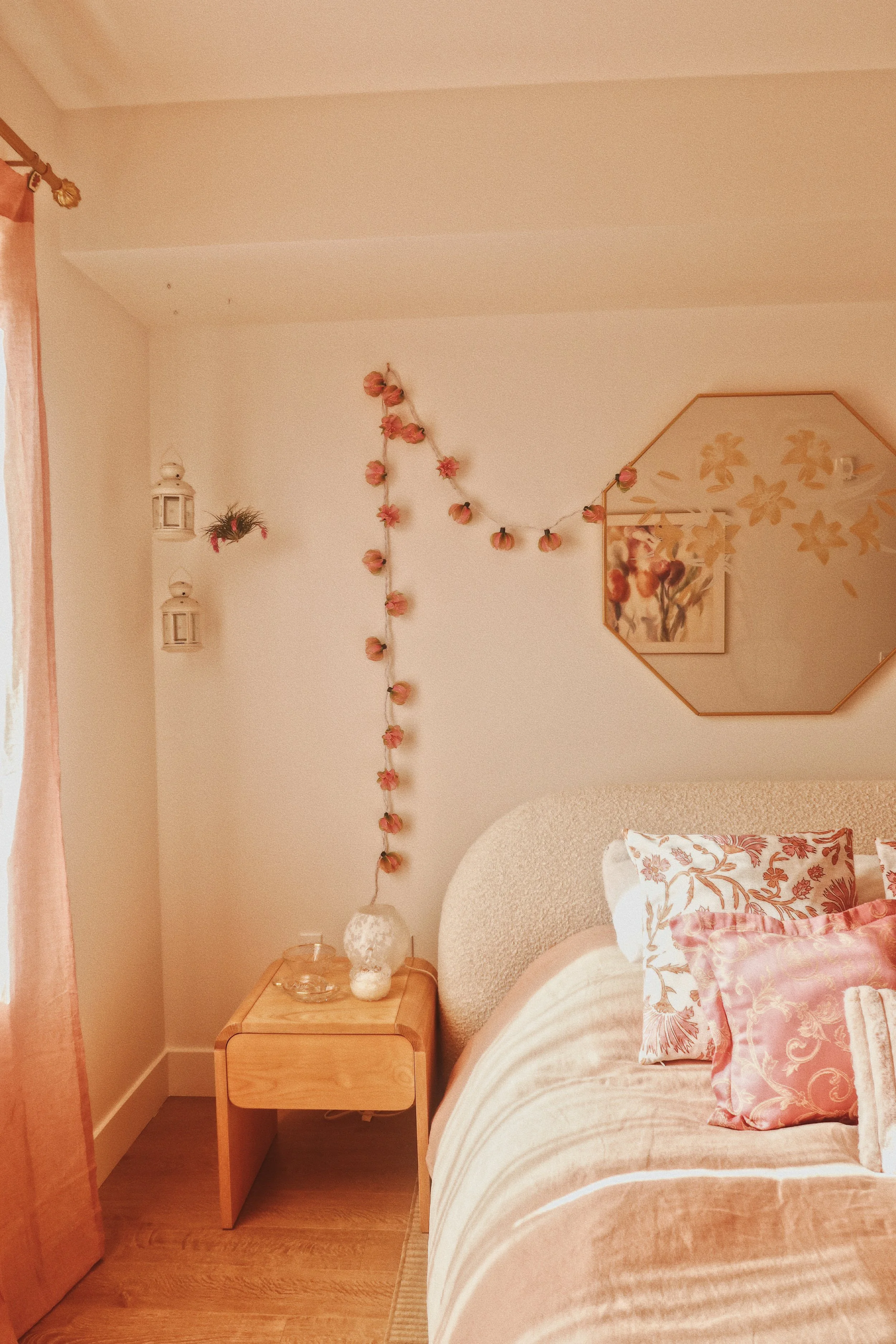 A cozy bedroom with a beige bed, pink and floral pillows, a wooden nightstand with a white vase and lamp, floral wall hanging, wall-mounted lanterns, and pink curtains, all creating a warm, inviting atmosphere.