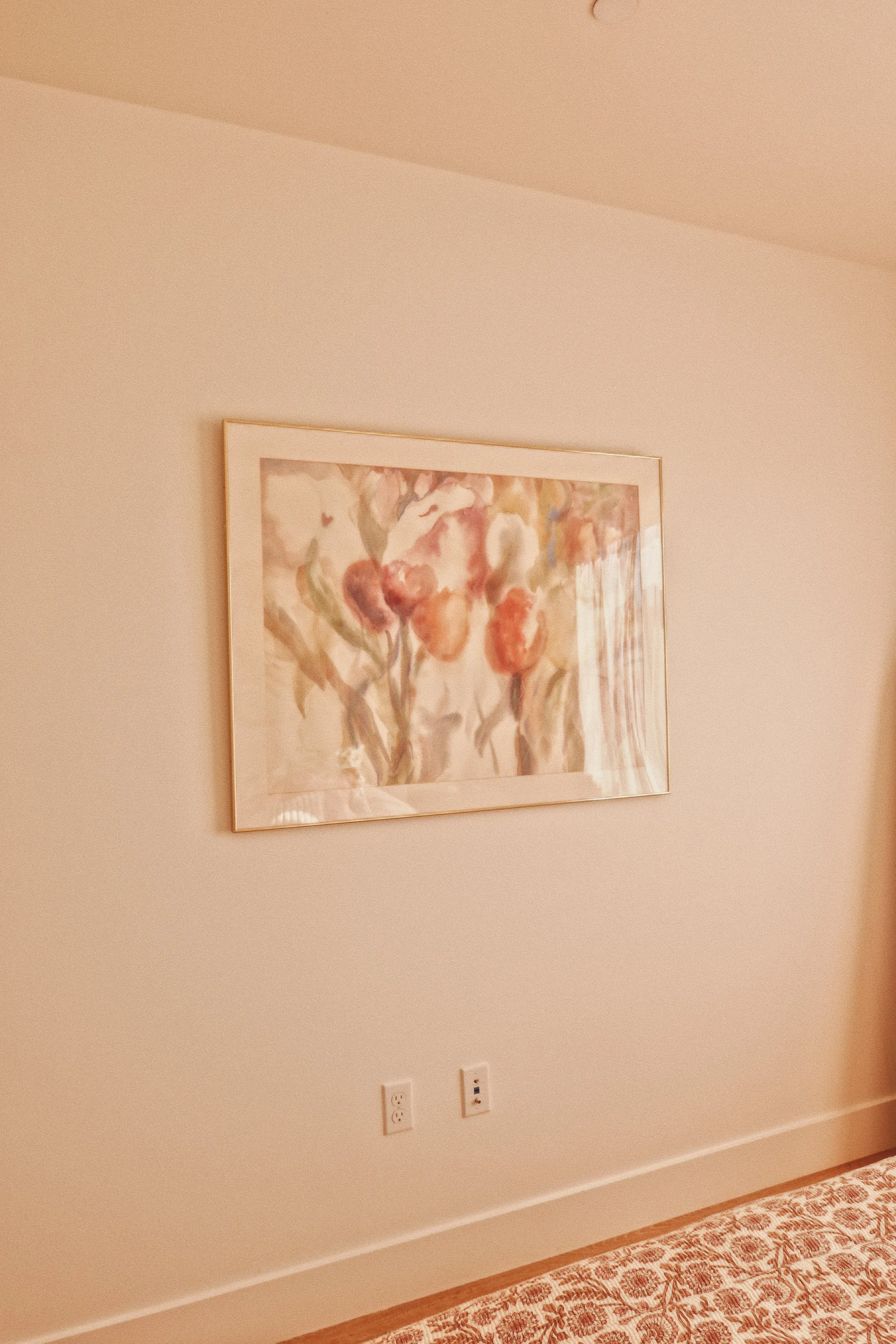 A corner of a bedroom with a framed watercolor painting of flowers on a beige wall, electrical outlets, a patterned bedspread, and a wooden baseboard.