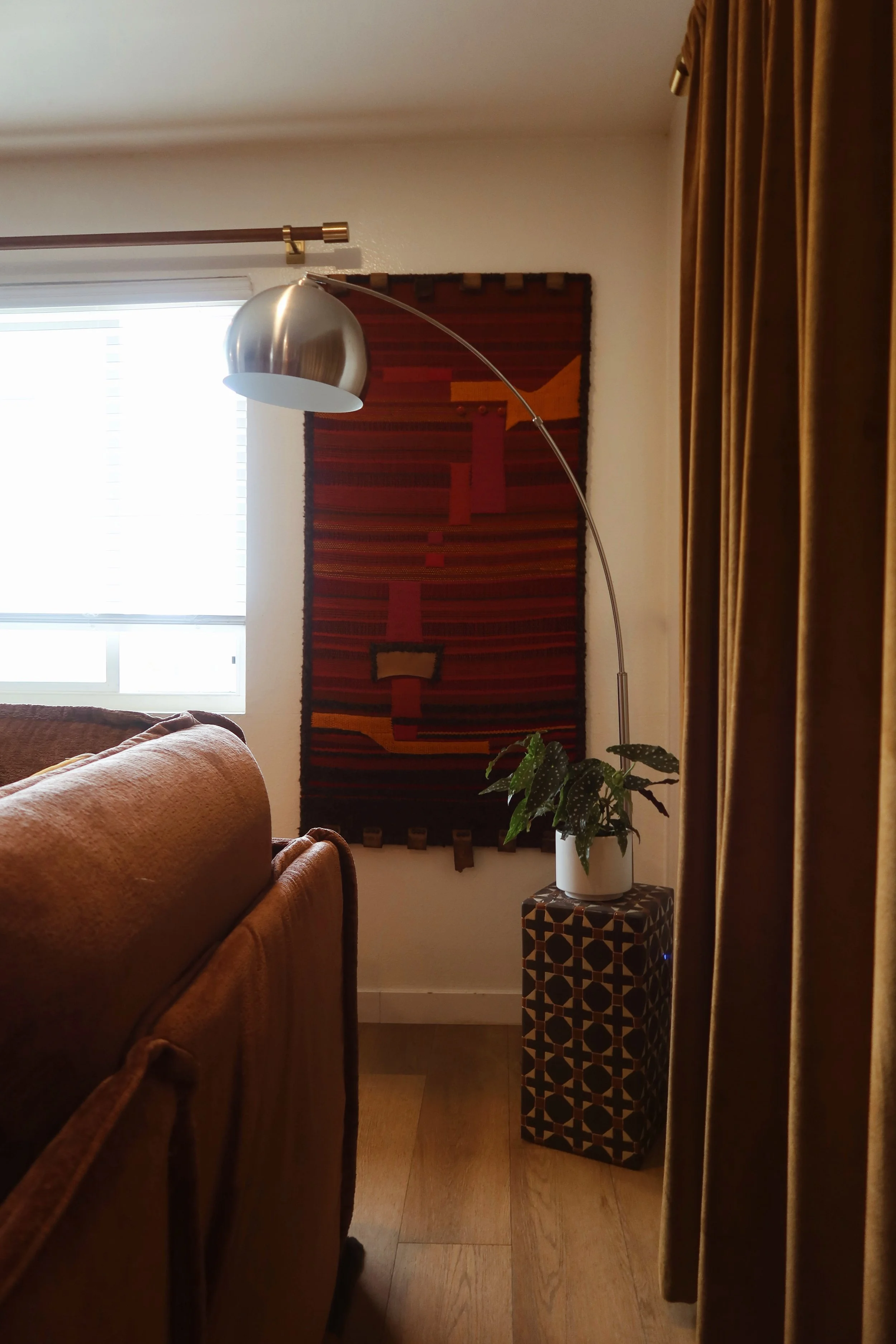 A cozy living room corner with a window, a mustard-colored sofa, a potted plant, a geometric side table, a floor lamp, a tapestry with red and orange patterns, and dark yellow curtains.