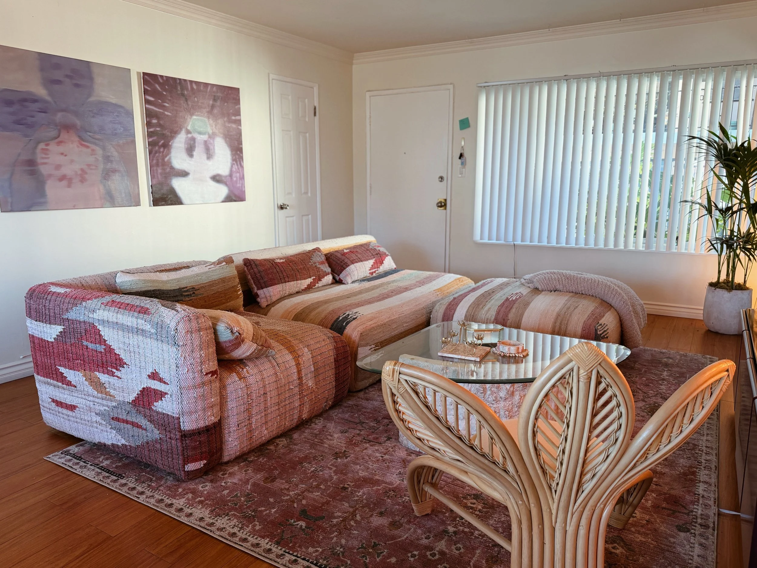 Living room featuring a wrapped fabric sofa with patterned cushions, a glass coffee table with decorative items, a large potted plant, and abstract artwork on the wall.