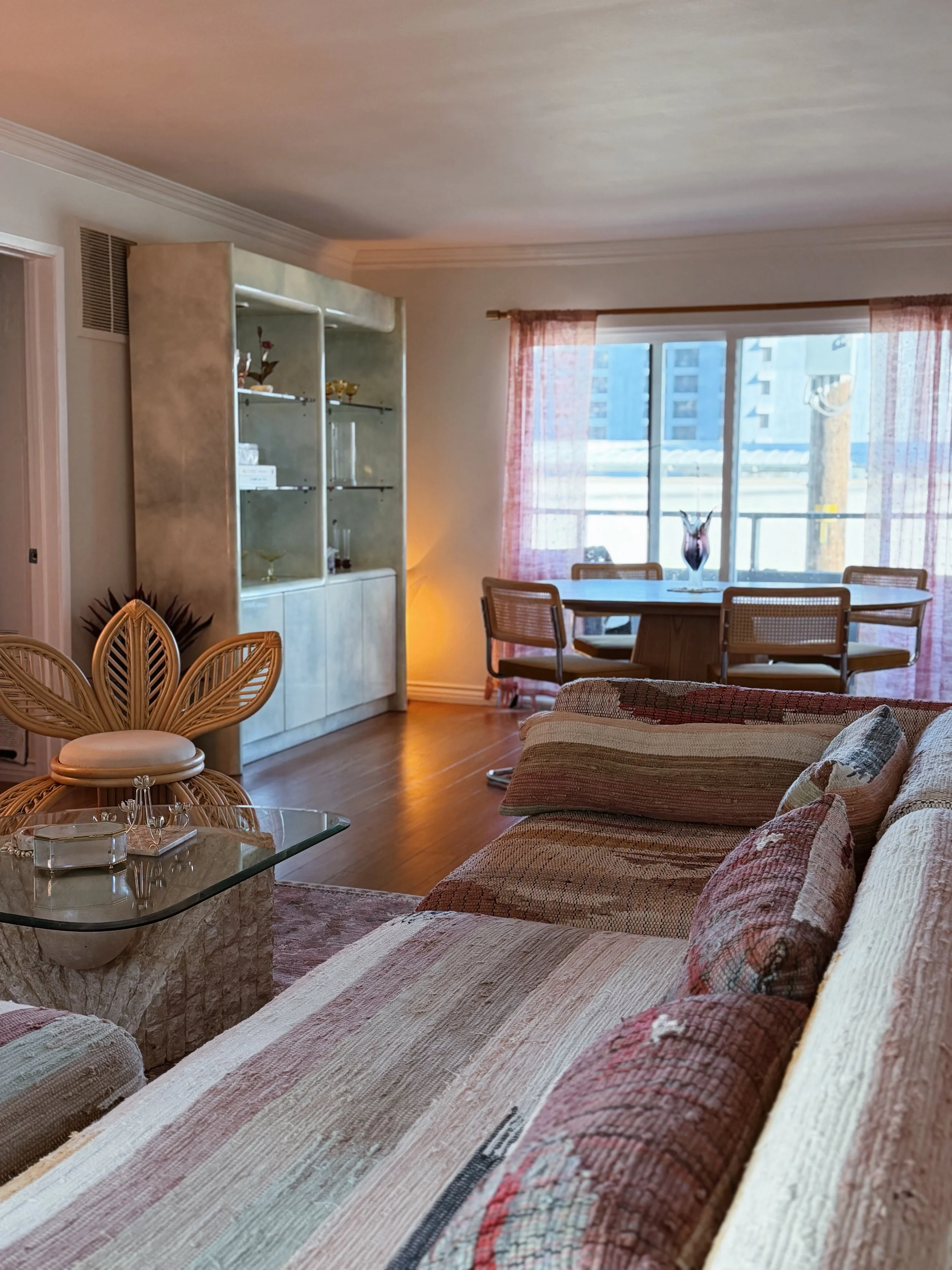 Living room with sofa, glass coffee table, dining area with chairs, large window with pink curtains, and built-in shelves.