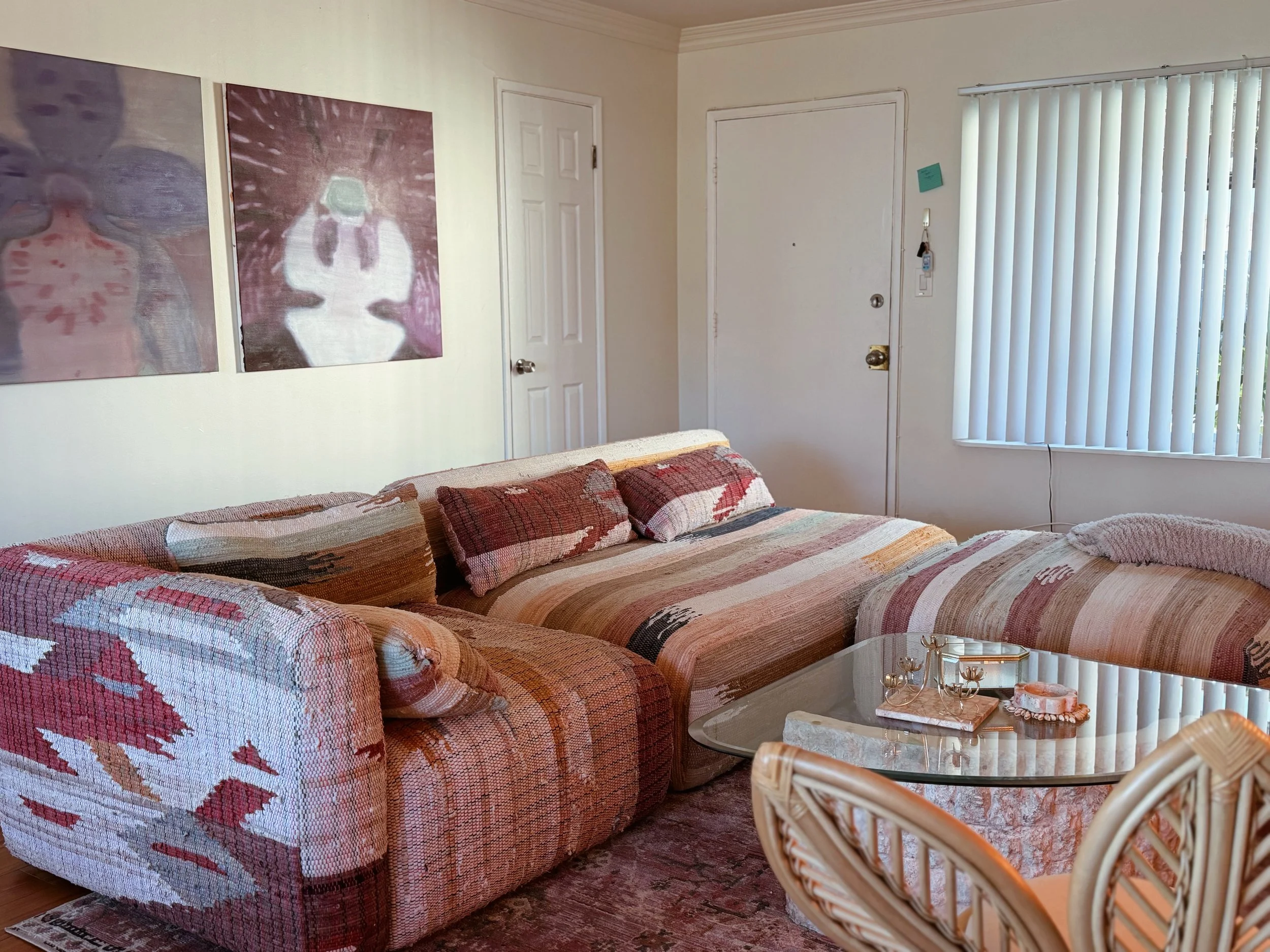 A living room with a multicolored woven fabric couch, a glass coffee table with decorative items, abstract artwork on the wall, a closed white door, and a window with vertical blinds.