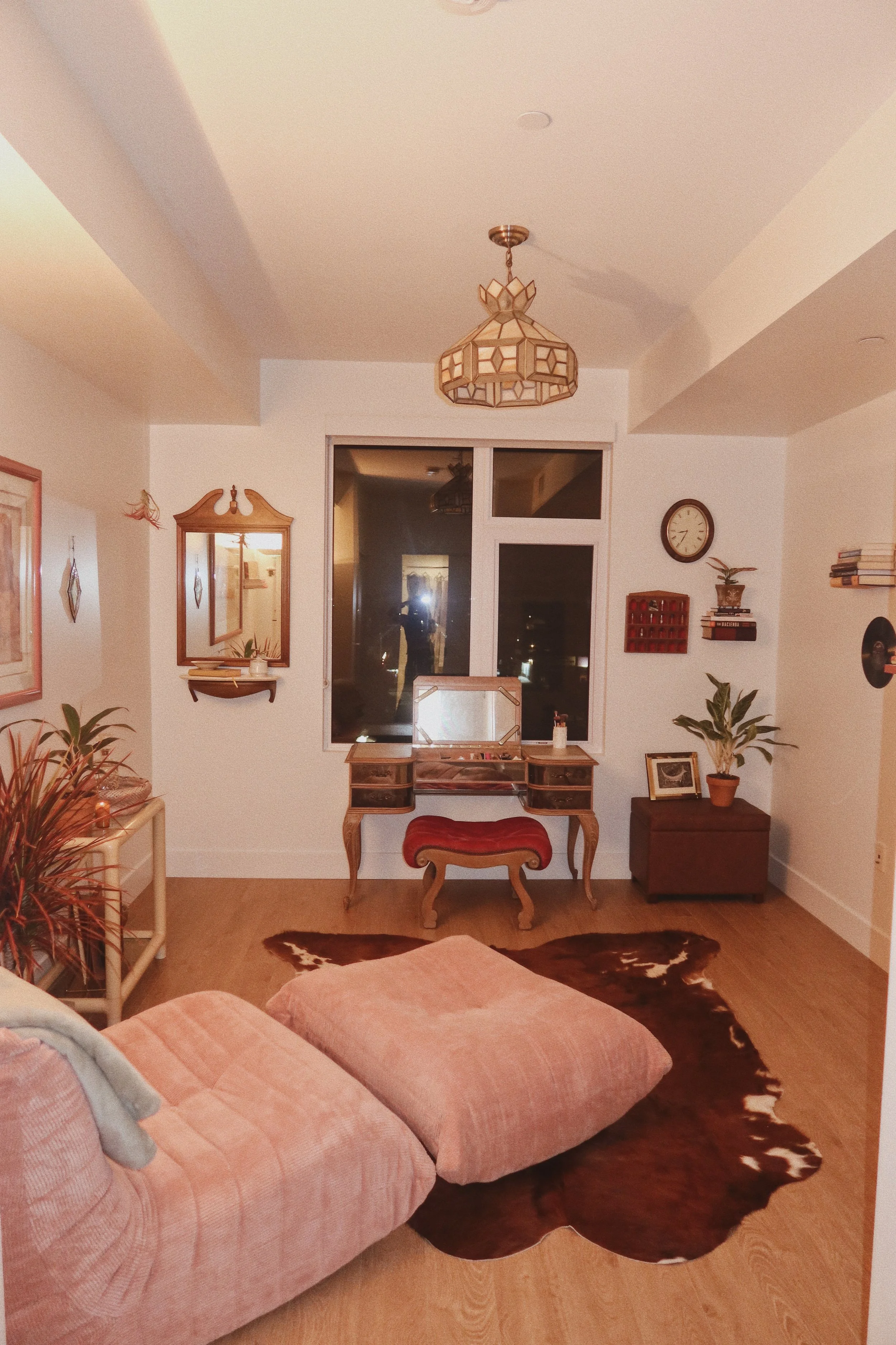 A cozy living room with a pink upholstered sofa, a cowhide rug, and a vintage wooden vanity with a mirror. The room has plants, framed pictures, and wall-mounted shelves with books and decor. A window shows a dark exterior night scene. A ceiling ligh