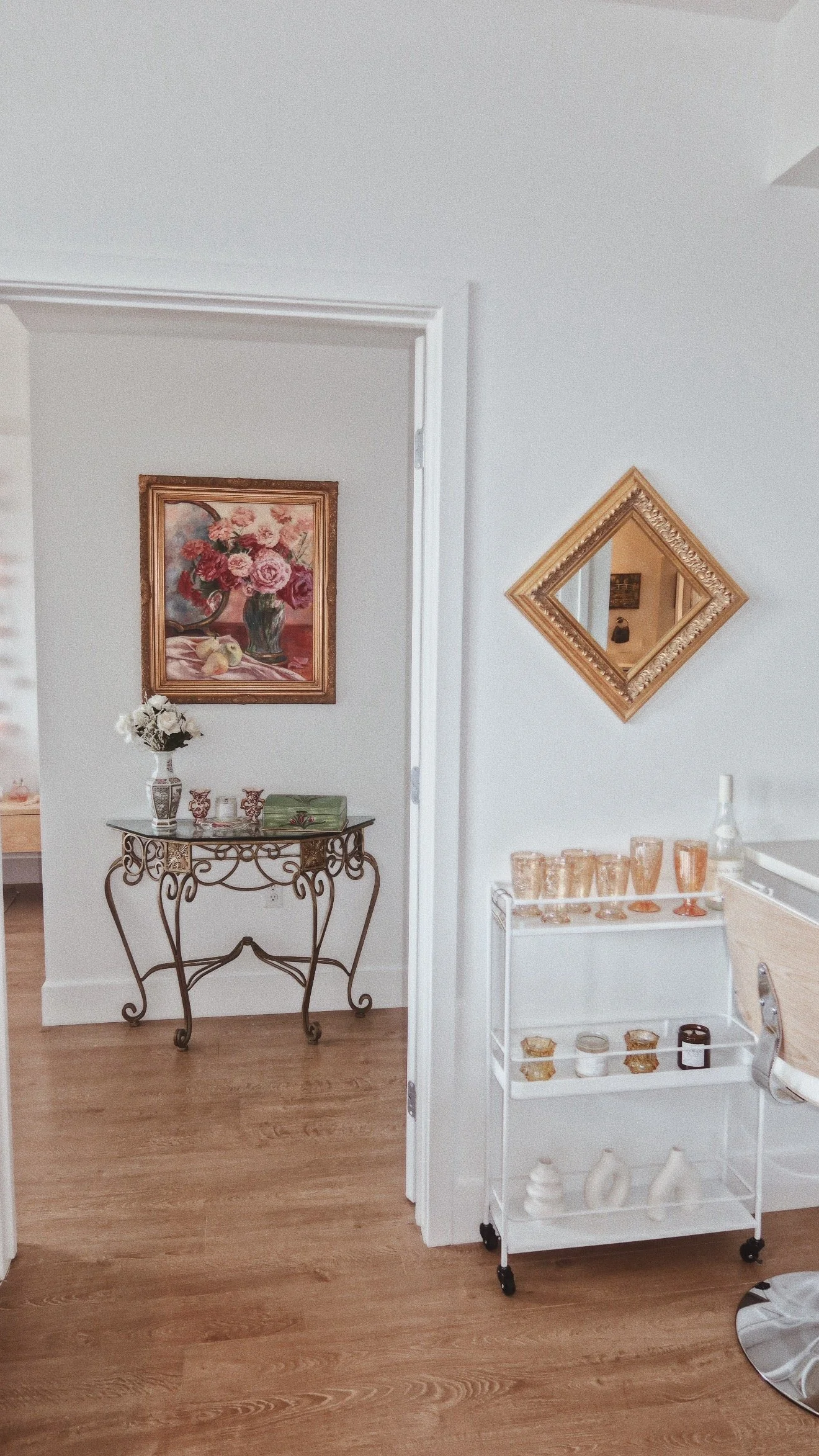 A corner of a room with a white wall, featuring a framed floral painting, a small decorative table with a vase of white flowers, and a metal stand with rose-colored glassware and white decorative items. A square mirror with a gold ornate frame hangs 