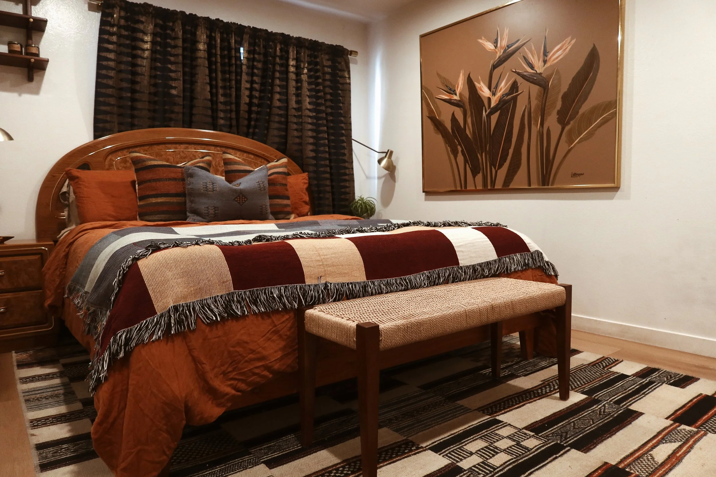 A bedroom with a wooden bed frame, orange and striped pillows, a blanket with red, black, gray, and white stripes, dark patterned curtains, a large framed floral painting, a small bench at the foot of the bed, a rug with black, white, and orange stri
