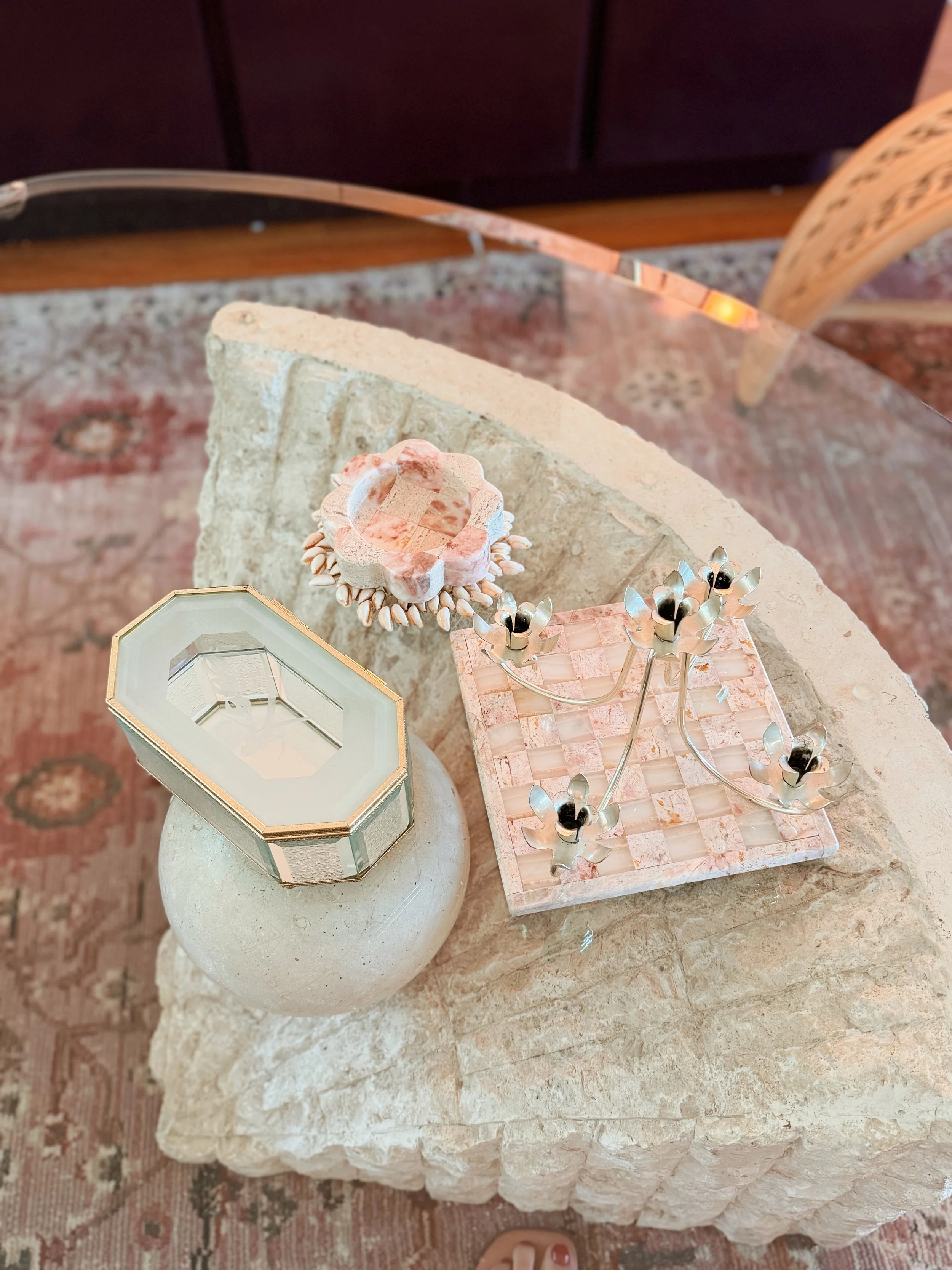 Decorative objects on a stone table, including a crystal candle holder, a pink marble ring with shells, a candelabrum with black candles, and a small mirror with a gold border.
