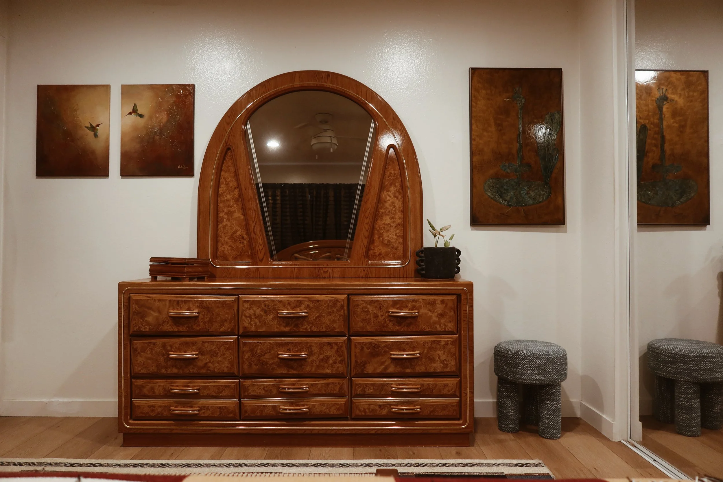 A wooden dresser with a large mirror on top, three drawers, and a small plant in a black pot on the right side, flanked by two patterned stool chairs. Three paintings of flowers hang on the wall above the dresser.