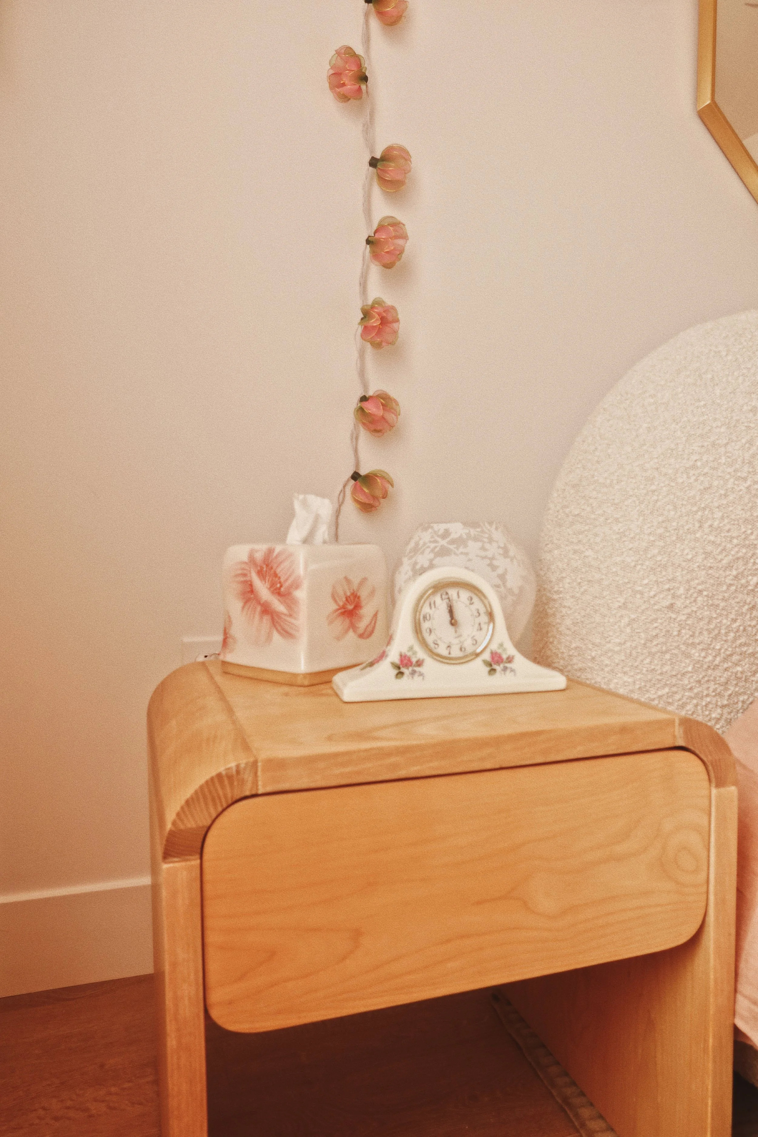 A wooden bedside table with a clock, a floral-patterned tissue box, and a white decorative vase with pink flowers, placed against a beige wall with a string of pink flowers hanging.