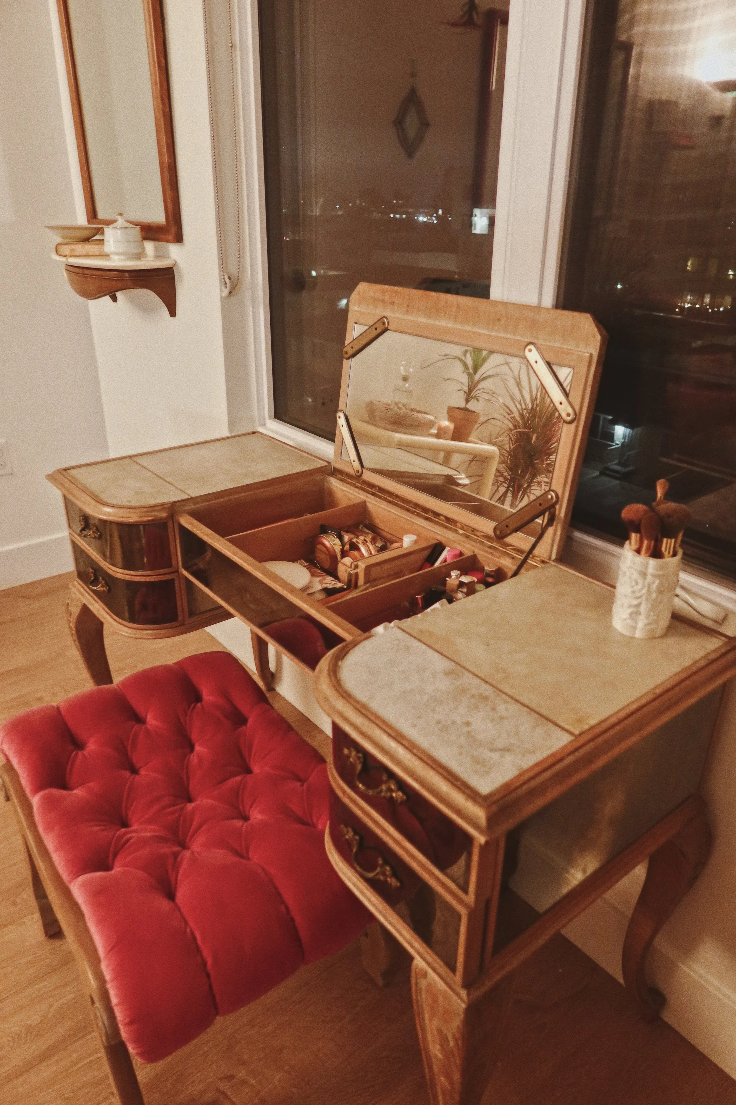 A vintage vanity table with a mirror and a drawer, adorned with makeup items, along with a red tufted upholstered chair, inside a cozy room with wooden flooring and a large window.