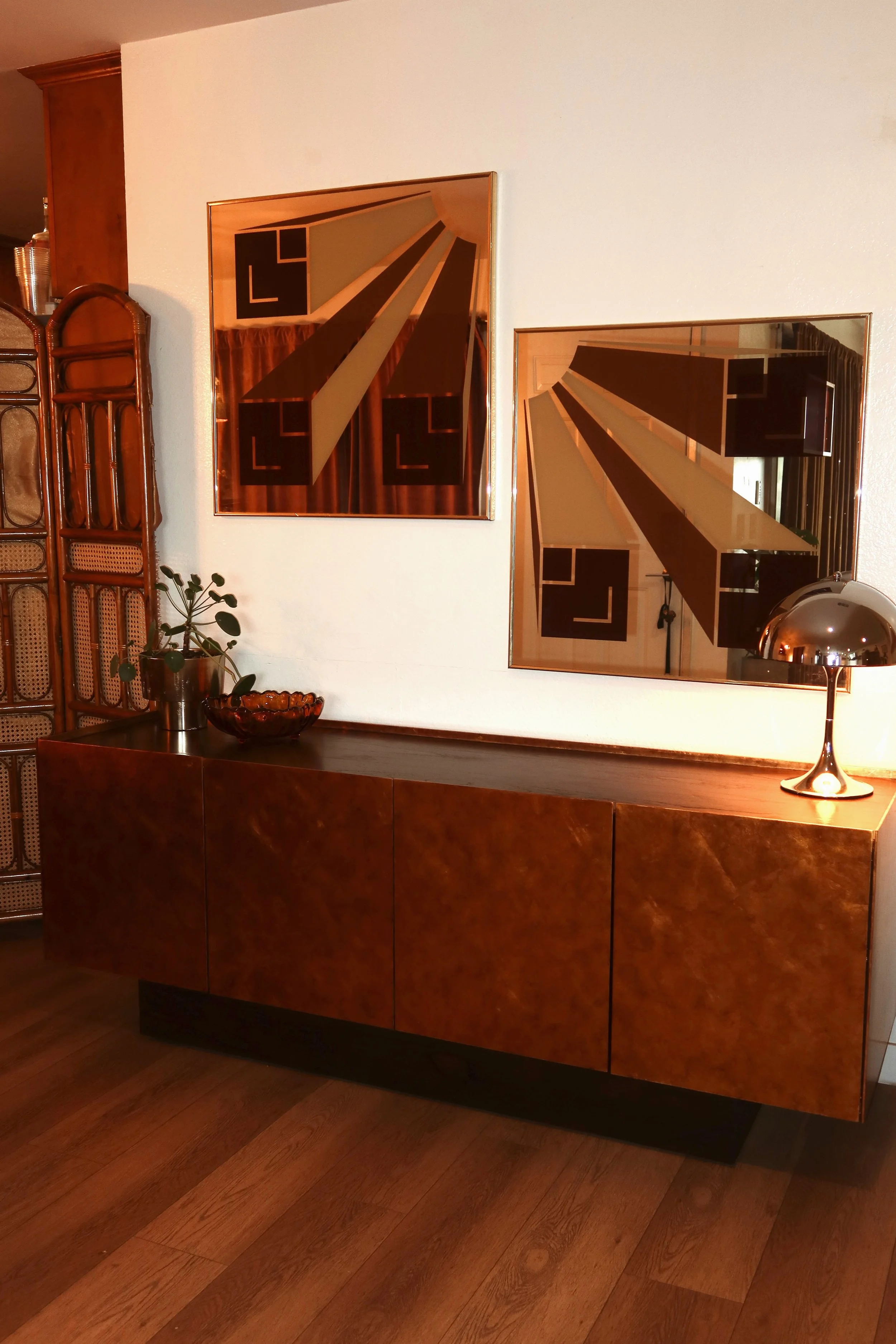 A wooden sideboard with a dark finish in a room with hardwood floors. Above it are two framed geometric abstract art pieces with brown, beige, black, and green colors. On top of the sideboard, there's a small potted plant, a decorative bowl, and a sh