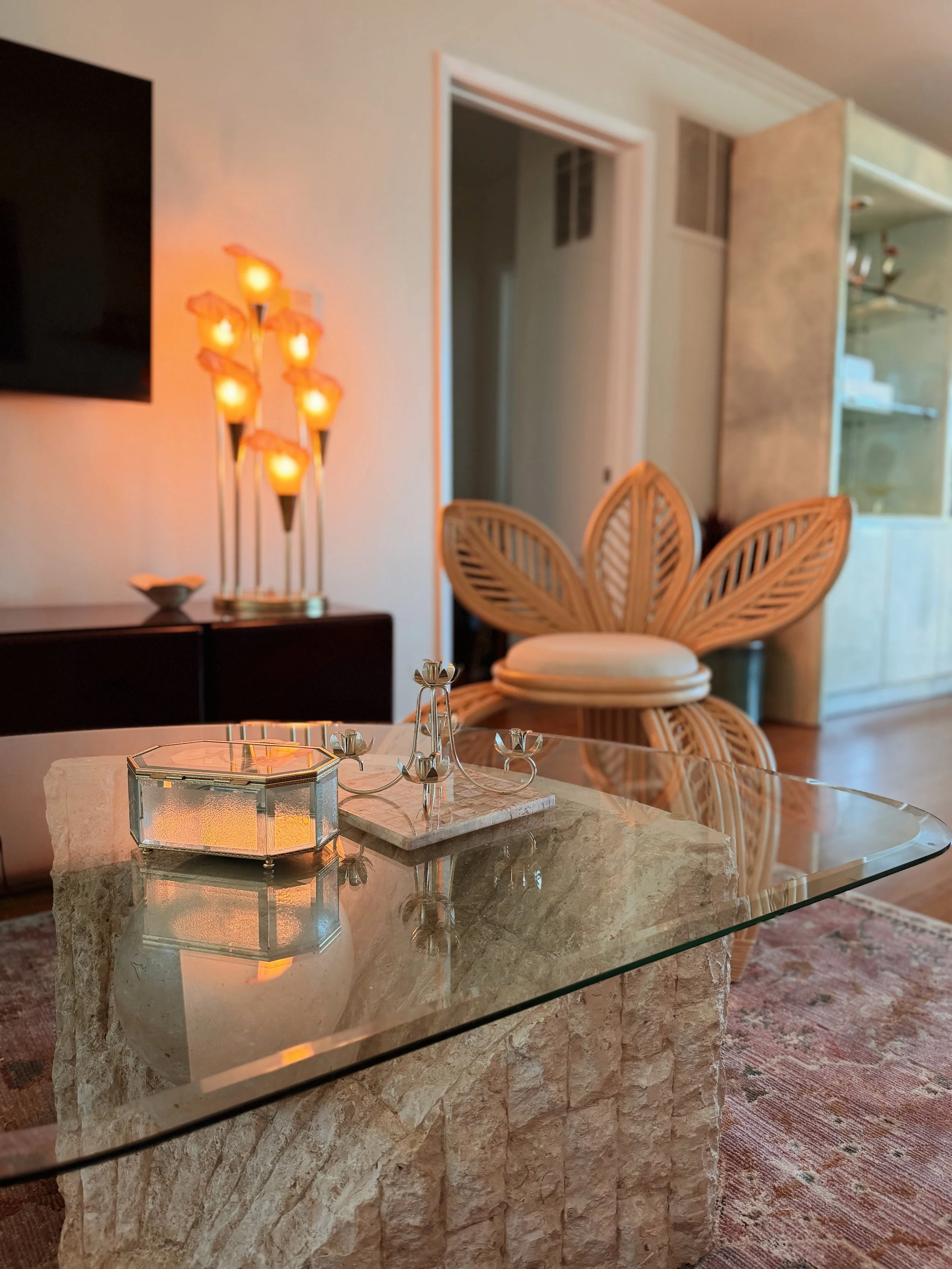 Close-up of a marble table with a silver candelabra and a decorative glass box in a living room. In the background, there are wooden chairs with woven backrests, a wall-mounted black TV, and a yellowish wall with modern lamp fixtures.