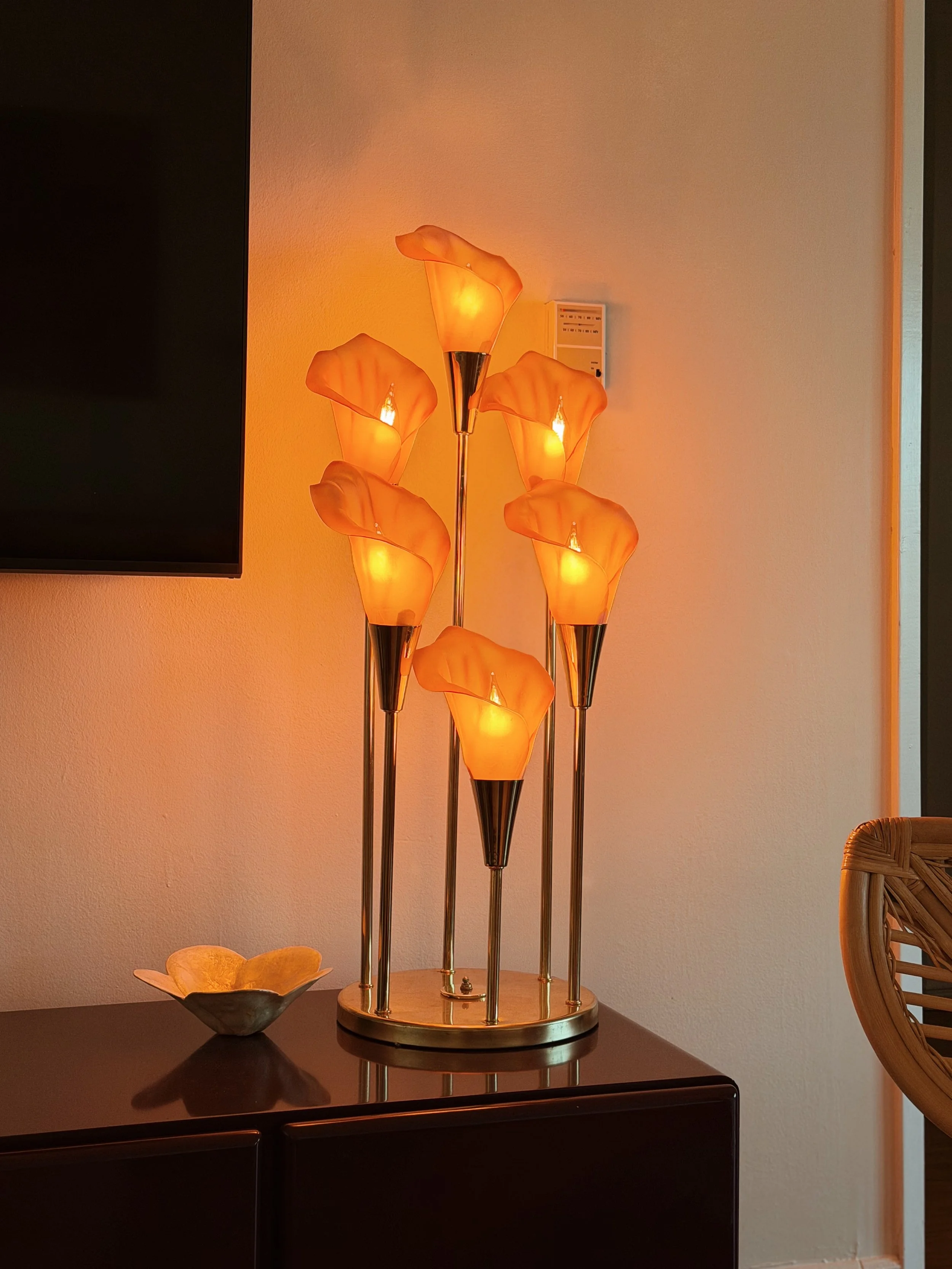 A modern floor lamp with five orange, flower-shaped glass shades on a brass stand, placed on a dark wooden table next to a bowl shaped like a flower.