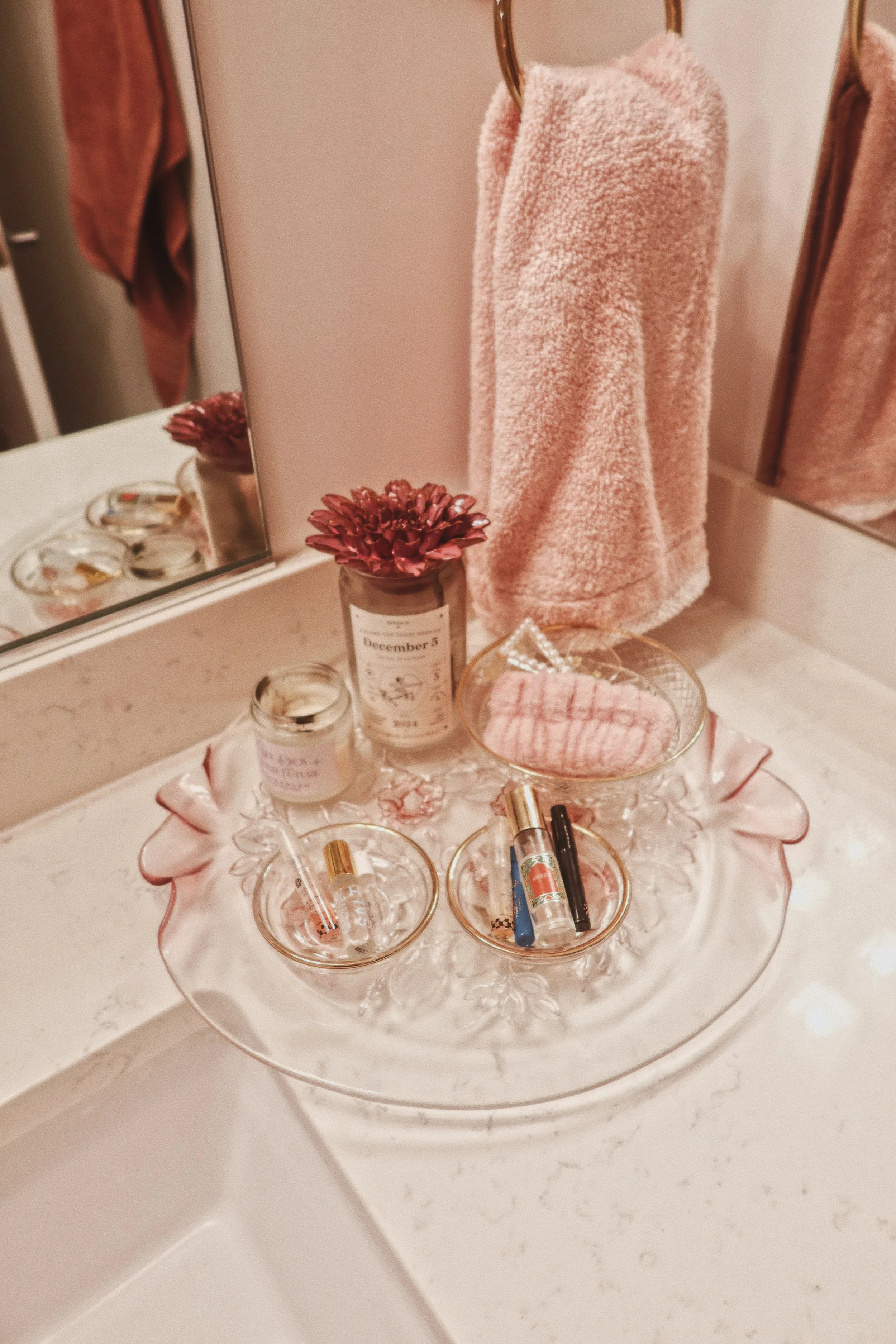 A glass tray with three small bottles, various makeup and lipstick products, and a pink towel.
