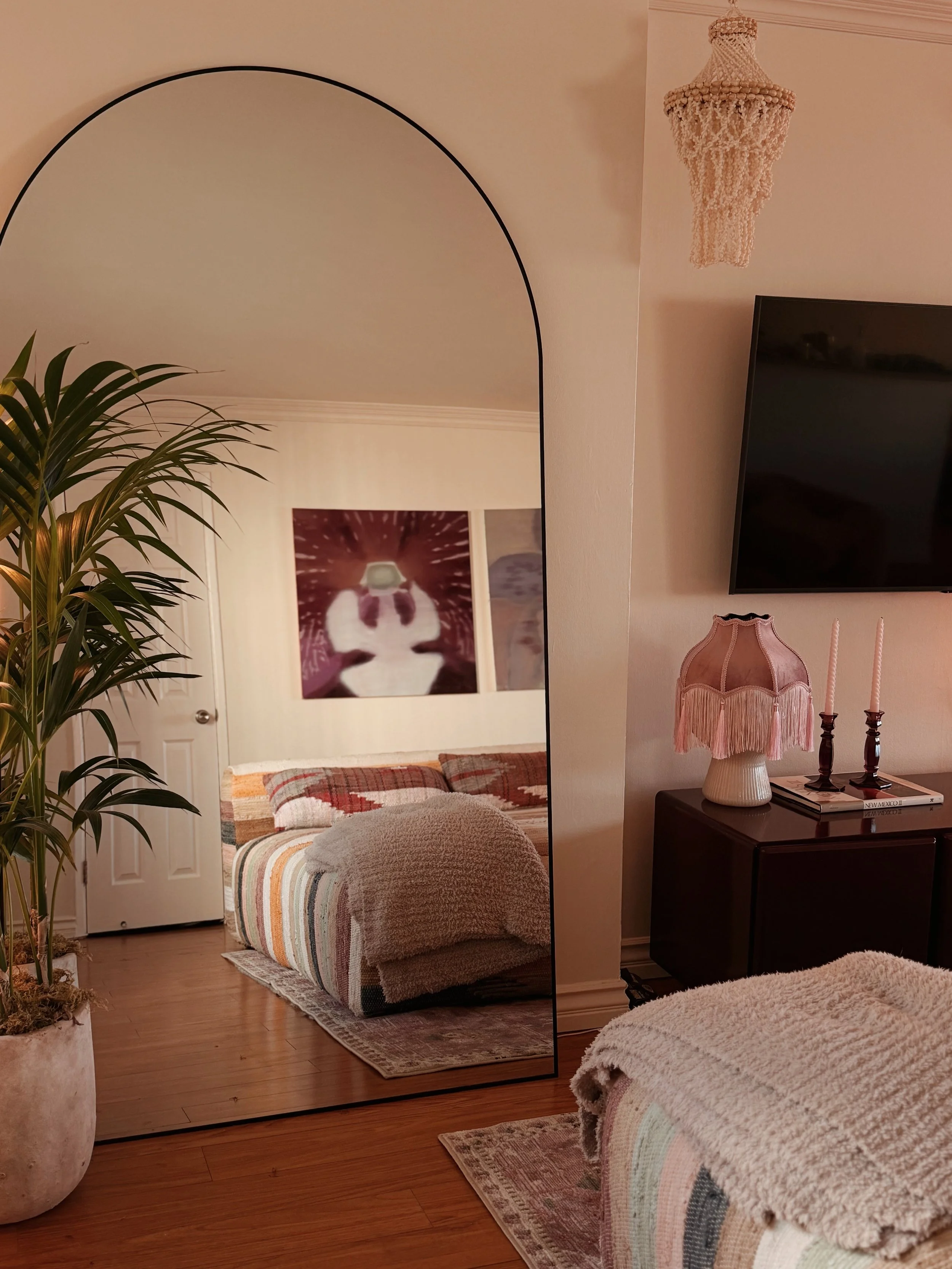 A bedroom with a large mirror reflecting a bed with a striped quilt and a basket. The room has hardwood floors, a potted plant, and a nightstand with a pink vase and candelabra.