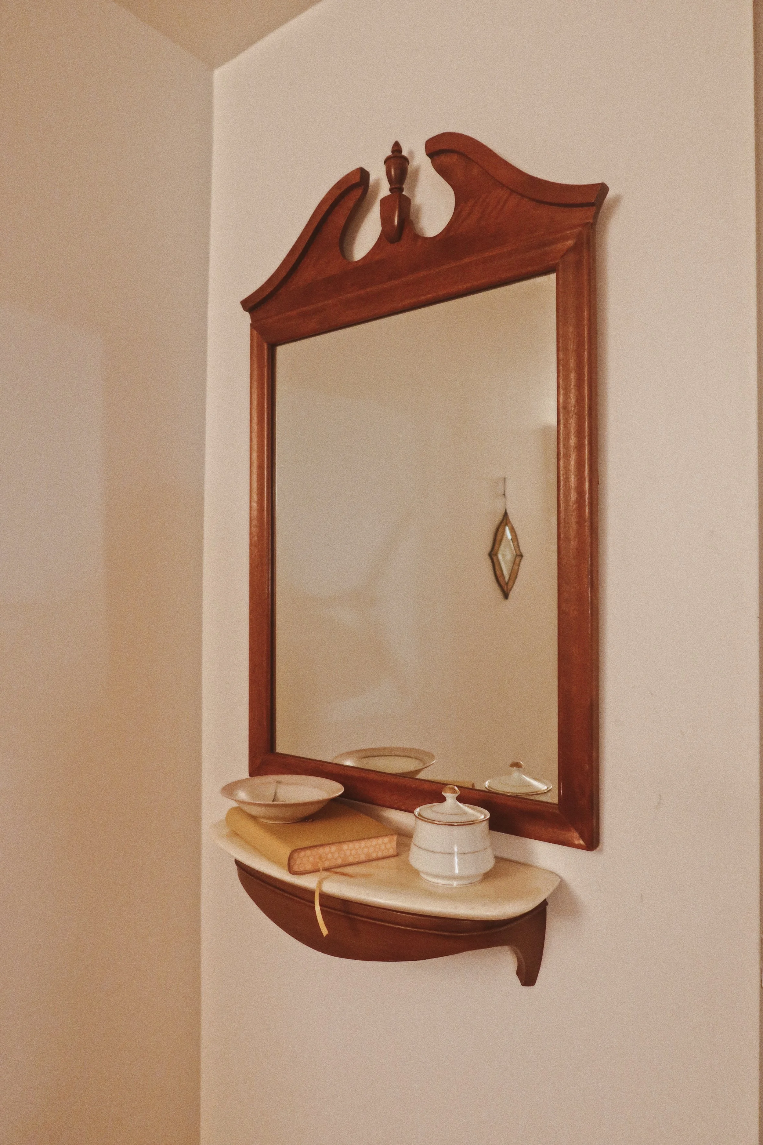 A wooden wall mirror with a shelf below, decorated with three bowls and a small white teapot, against a plain beige wall.