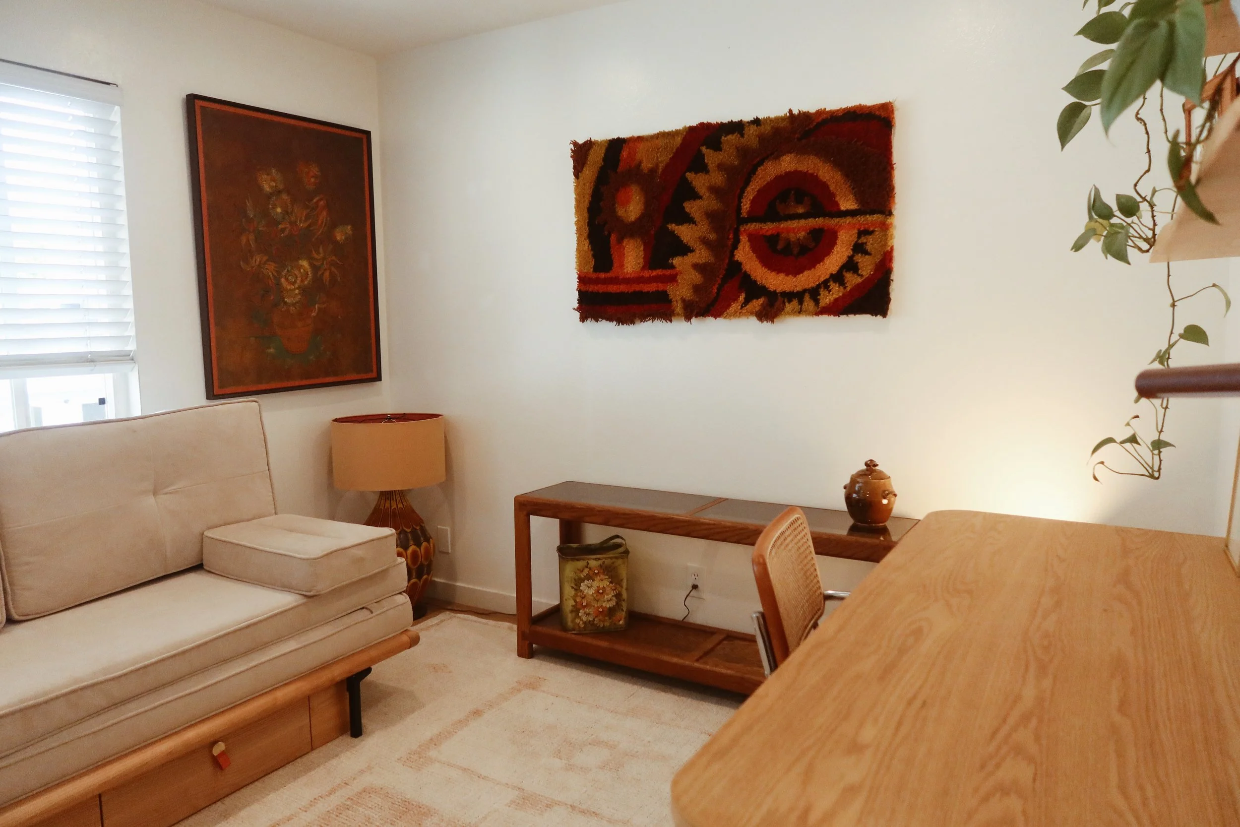 Living room with beige sofa, wooden coffee table, wall art, and a striped rug. There is a window with blinds on the left, a table with a plant and decorative vase, and a colorful abstract wall hanging.