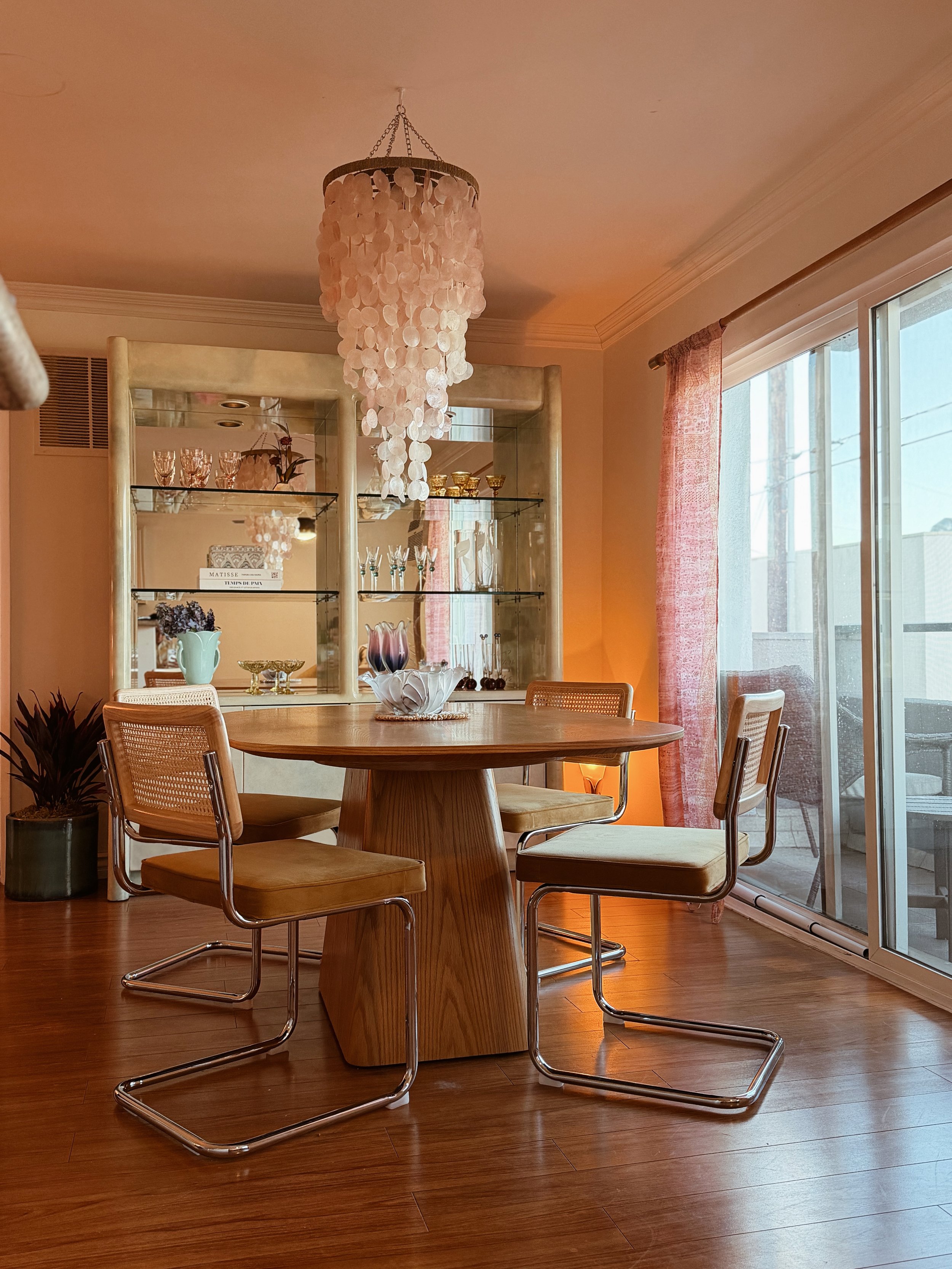 A dining room with a round wooden table surrounded by four modern chairs with beige cushions and chrome legs. A shell-shaped vase with purple flowers sits in the center of the table. A glass display cabinet with glassware and decorative items is in t