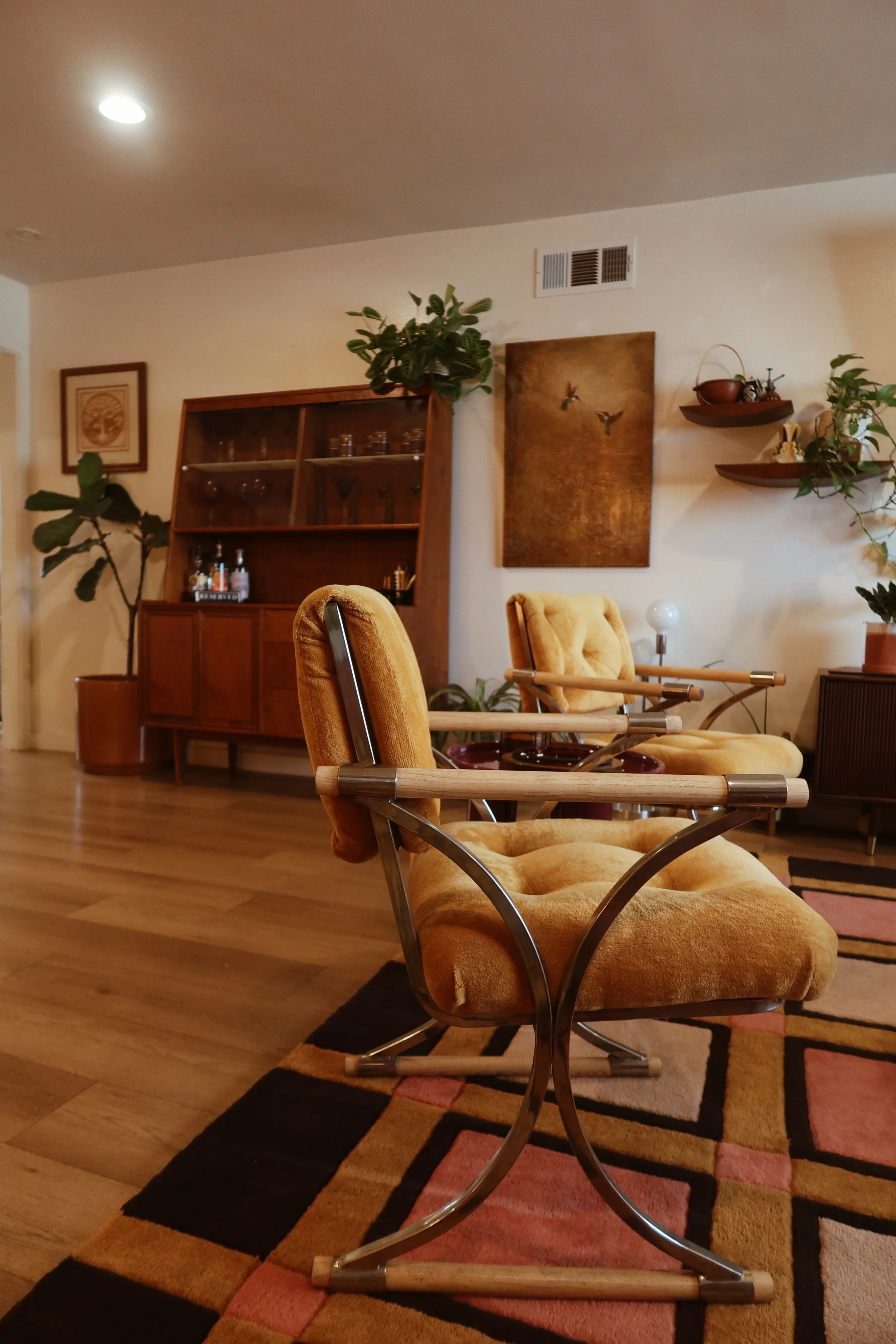 Living room with beige velvet lounge chair, wooden floor, Persian-style rug, wooden cabinet with glassware, framed artwork, plants, and decorative wall art.