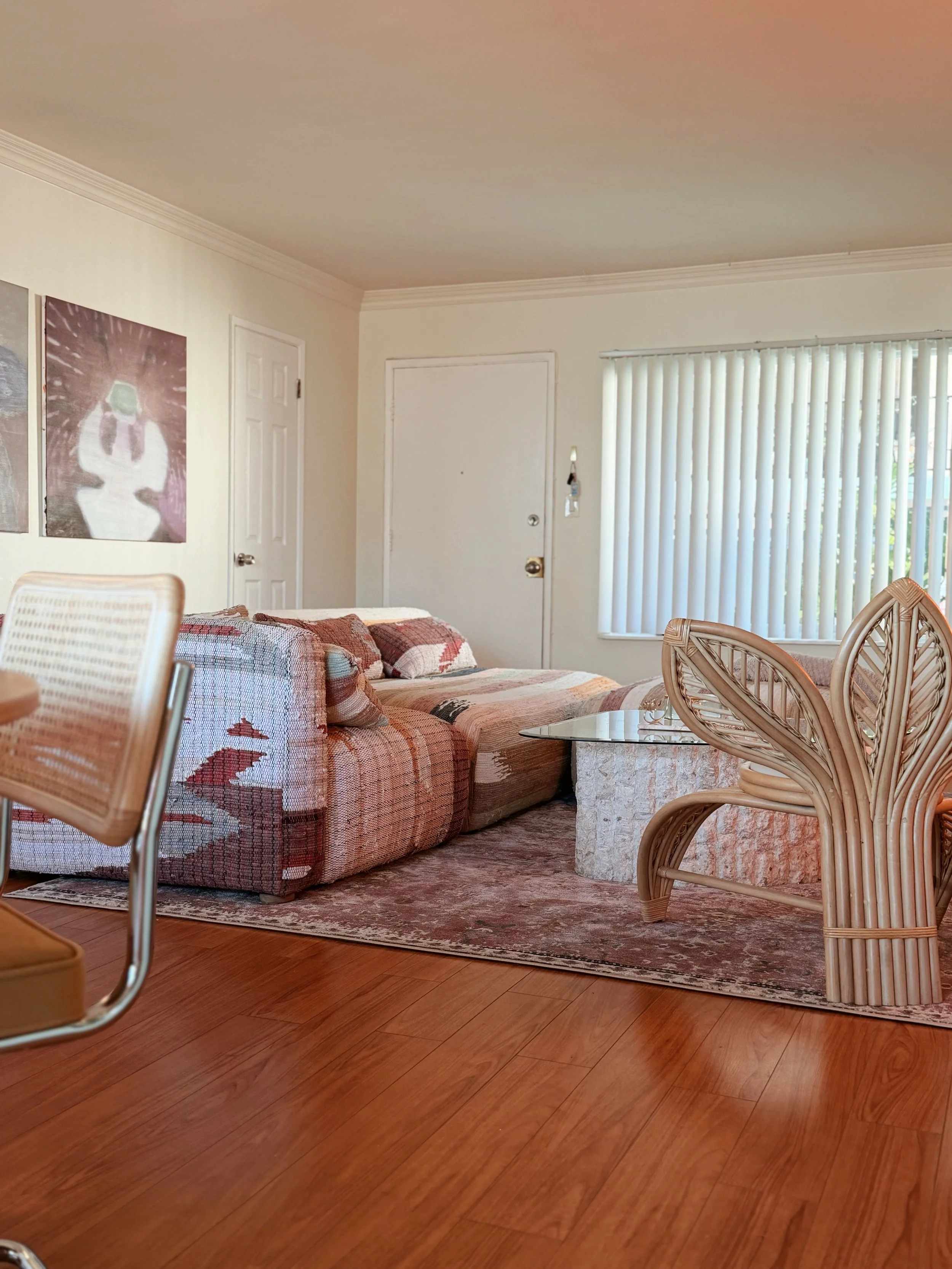 Living room with a plaid sofa, rattan chairs, glass coffee table, wood flooring, and vertical blinds covering a window.