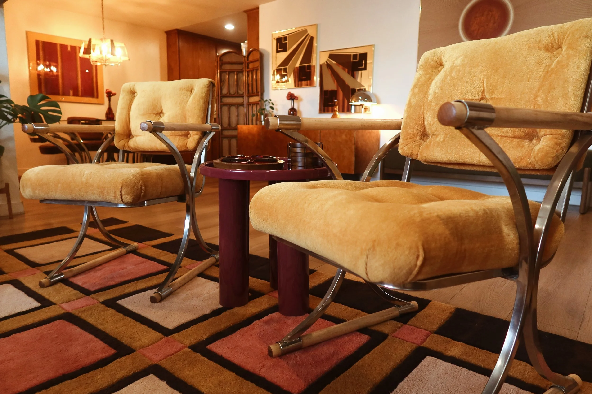 Living room with four yellow upholstered chairs with metal and wood armrests, a patterned area rug, and wall decorations including mirrors and paintings. There are also plants, a side table, and warm ambient lighting.