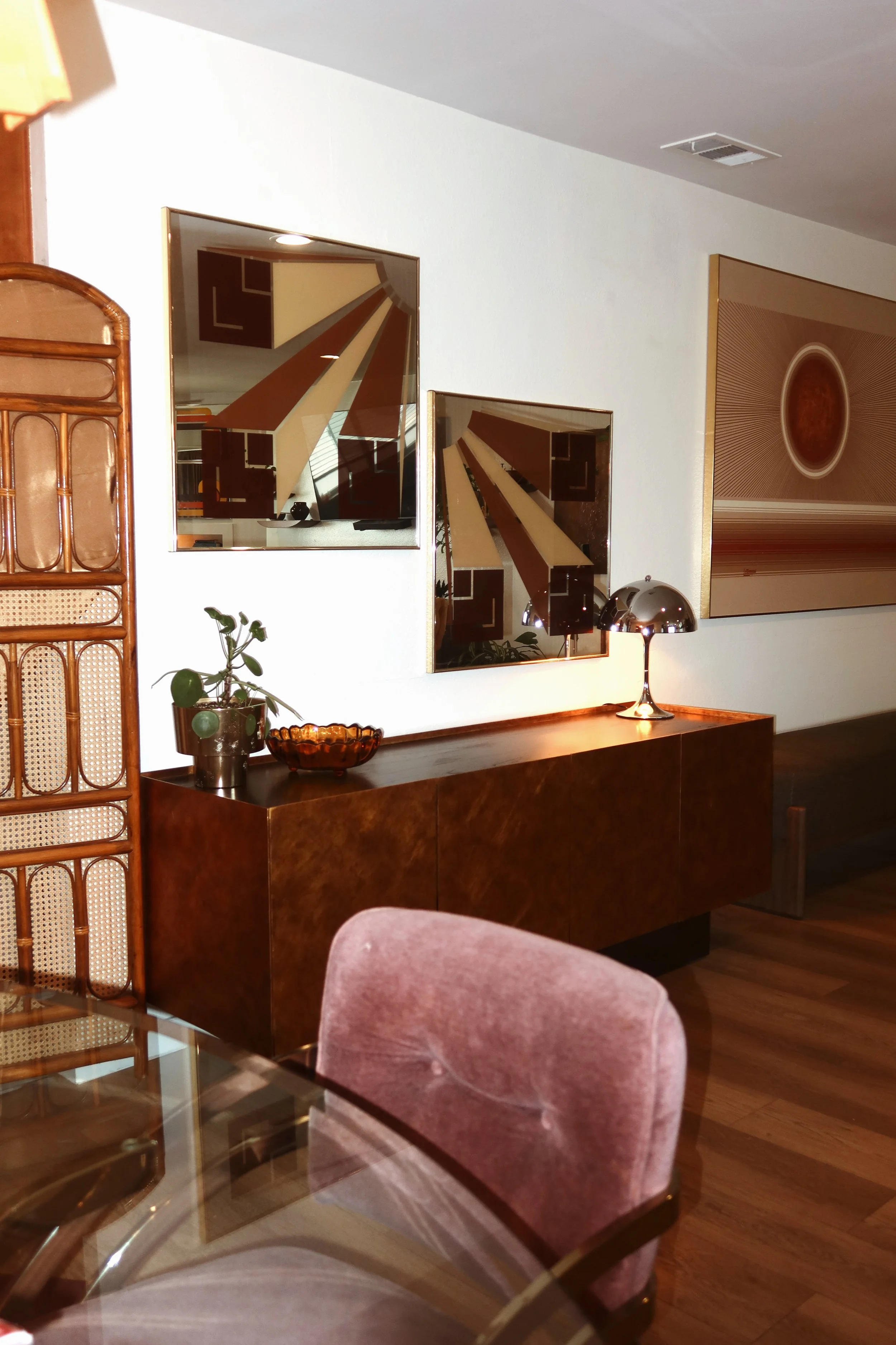 Living room with a wooden sideboard, two mirrors reflecting the ceiling, a lamp, a potted plant, a bowl, and a large artwork with a circular pattern on the wall.