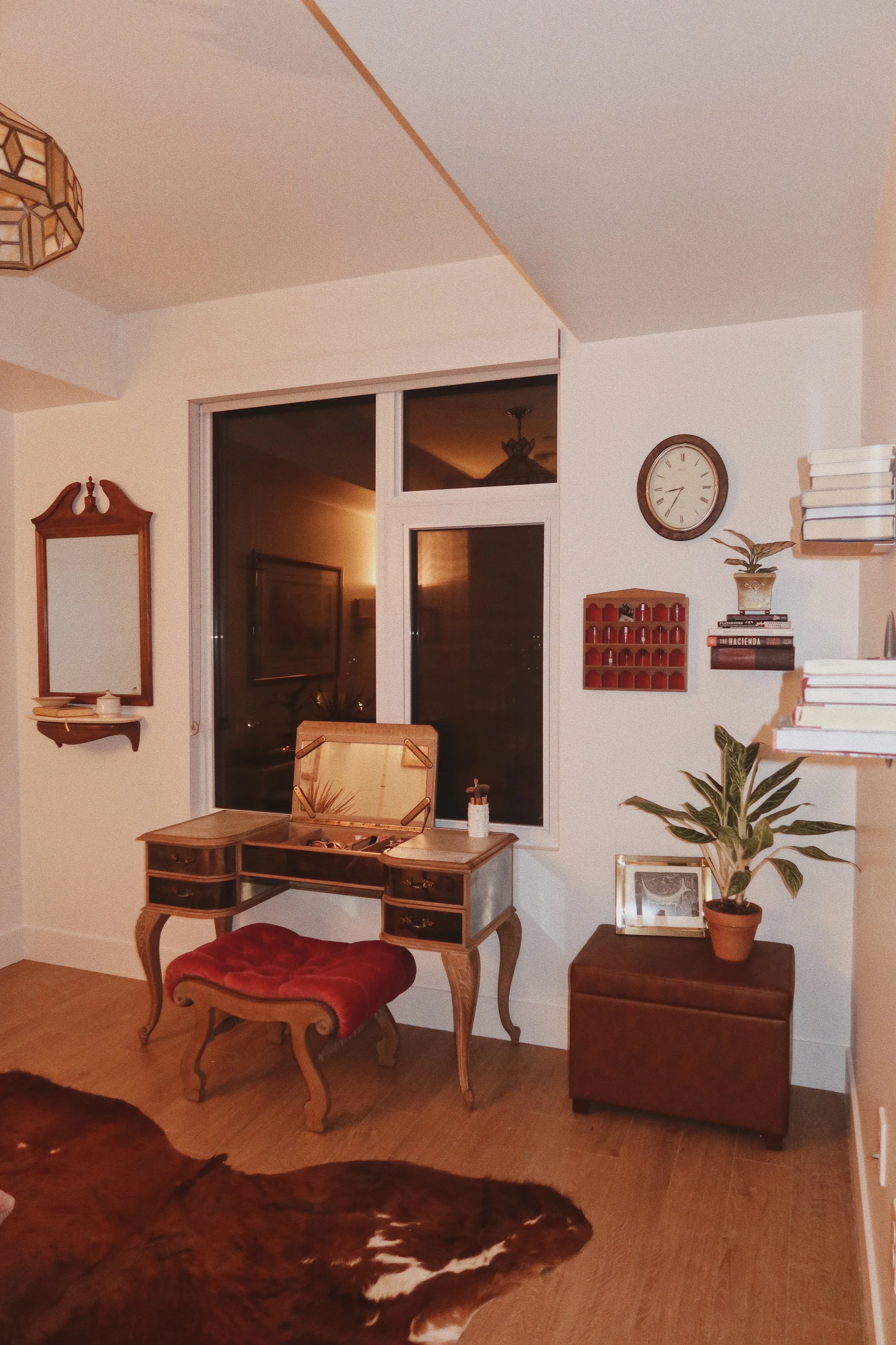 Living room corner with a vintage writing desk and red velvet stool, wall clock, bookshelf with books, potted plant, and window with a reflection of a ceiling light