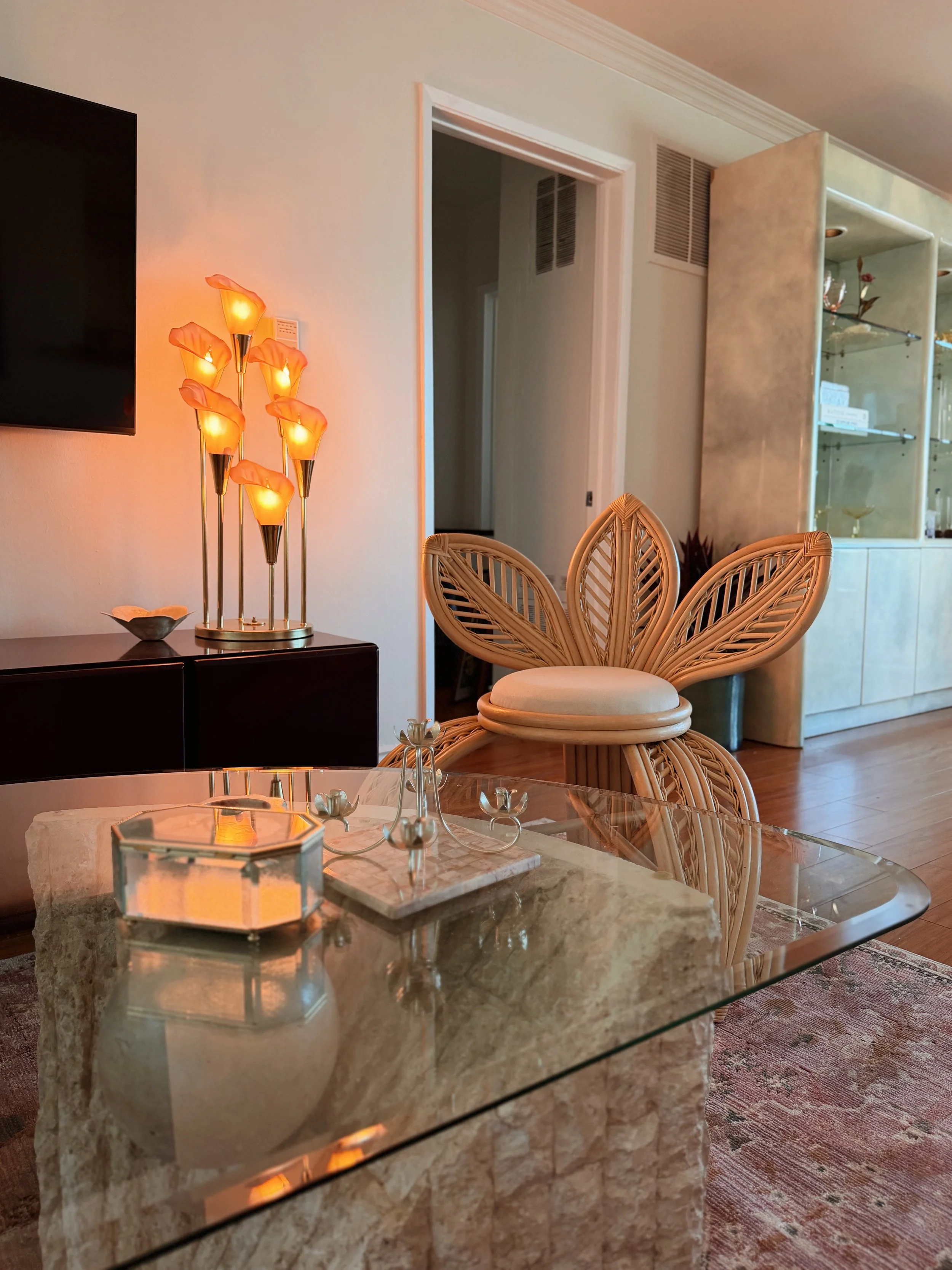 Living room with a glass coffee table, a rattan chair, a floor lamp with flower-shaped lights, a wall-mounted TV, and a glass display cabinet with decorative items.