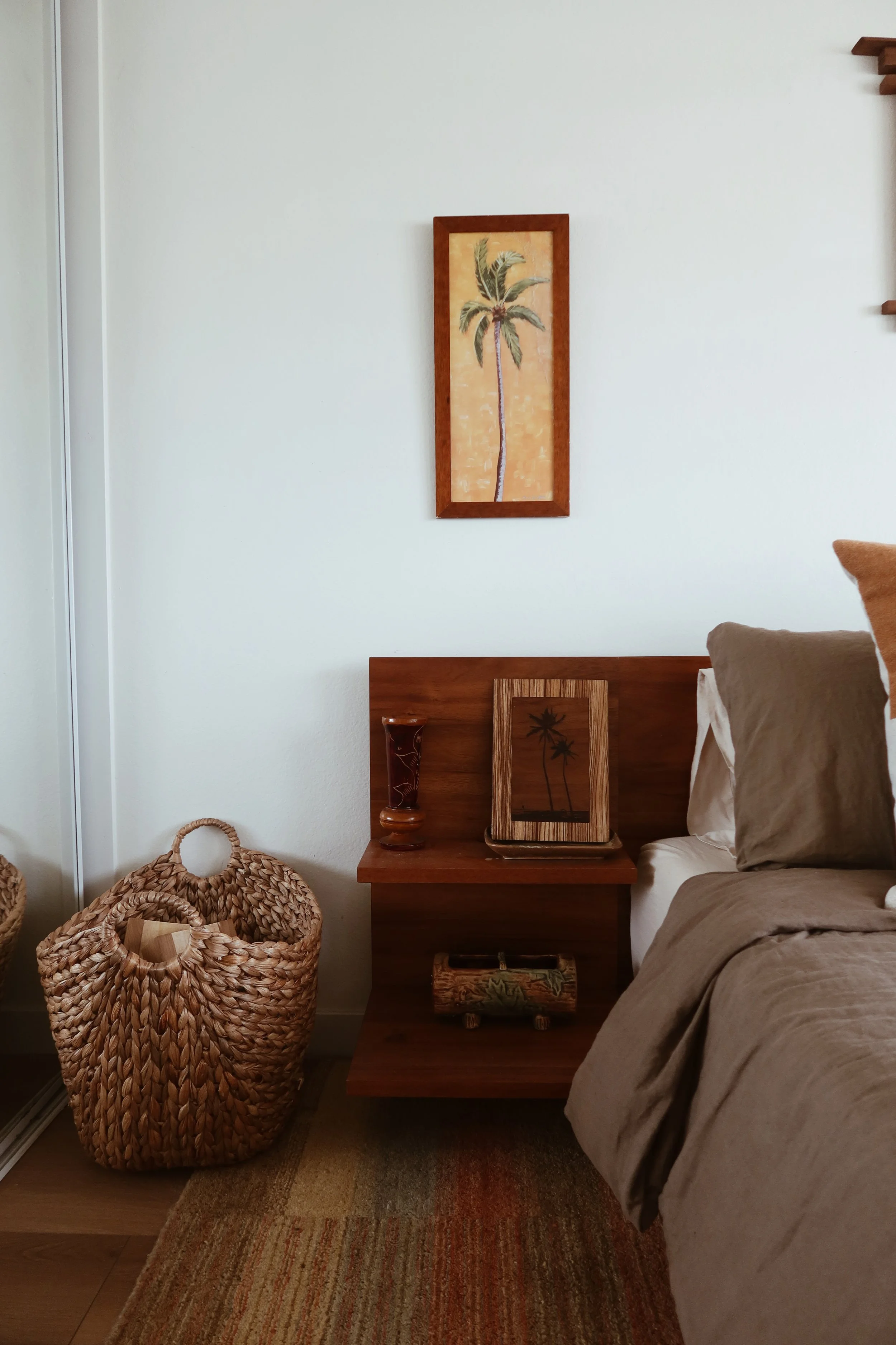 A bedroom corner with a woven basket, wooden nightstand with tropical-themed decor, a bed with beige bedding, and tropical wall art featuring a palm tree.